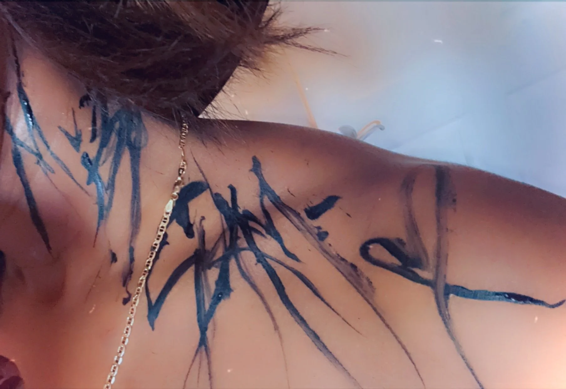 Close-up of a person's shoulder and chest with black ink tattoo art, gold chains, and long brown hair.