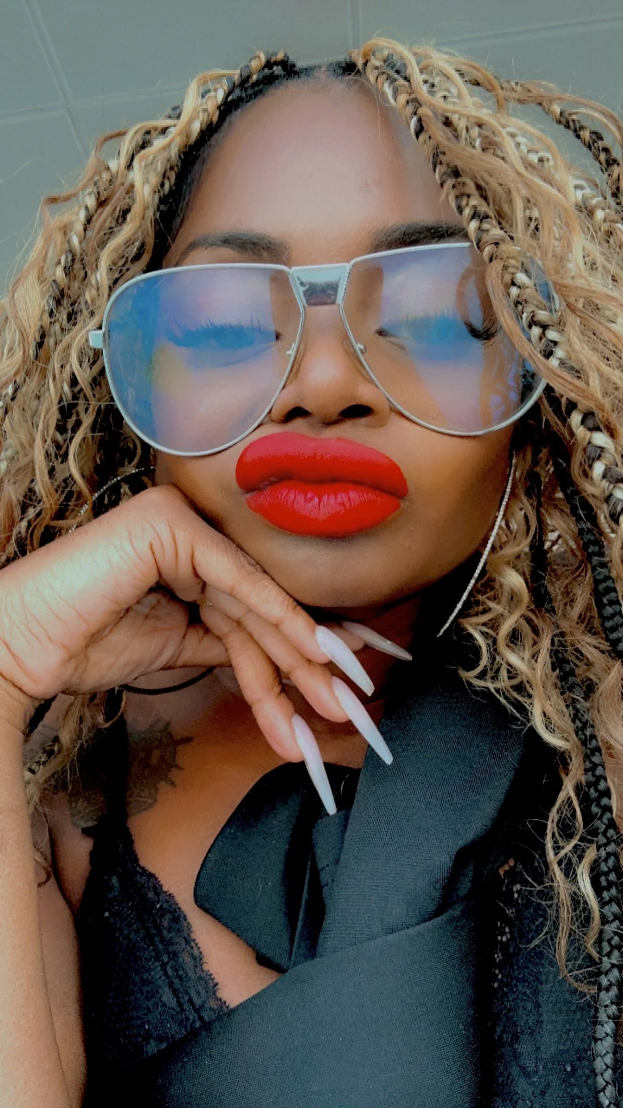 A woman with curly, blonde-highlighted hair wearing large reflective sunglasses with blue-tinted lenses and bright red lipstick, resting her chin on her hand, dressed in black.