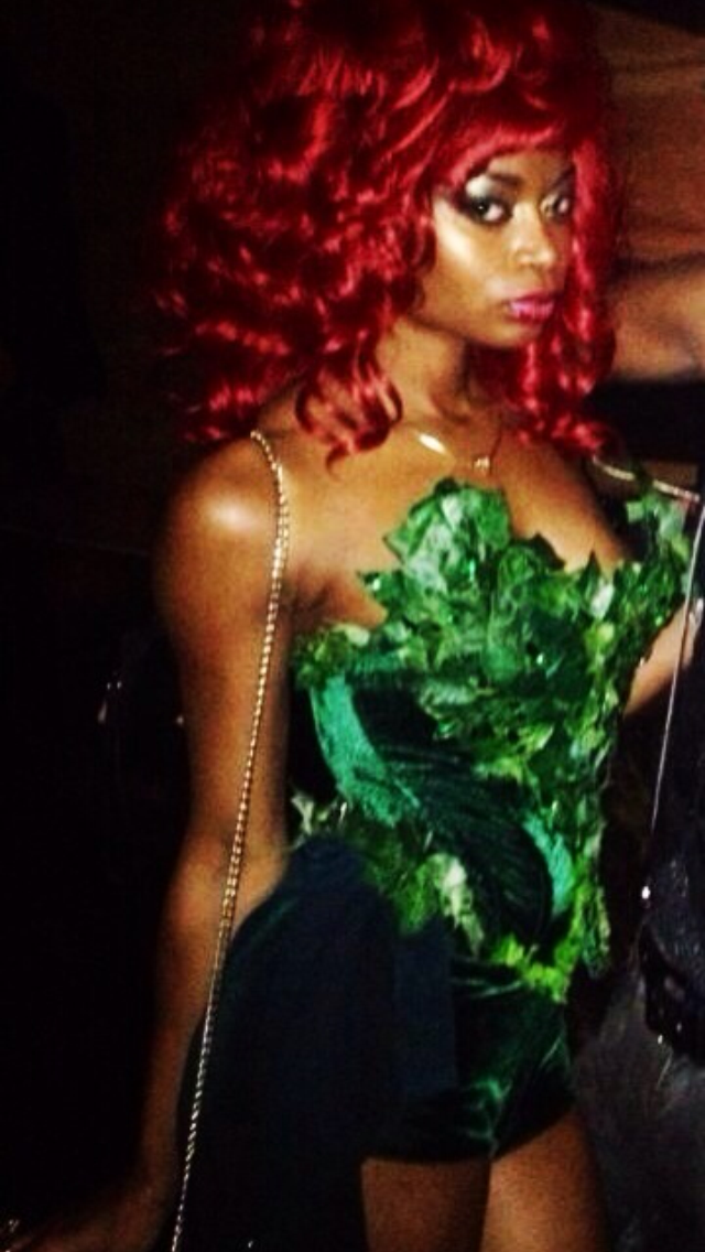 Woman with bright red, curly hair wearing a green leafy patterned strapless top, with a chain purse strap over her shoulder, looking to the side in a dimly lit setting.