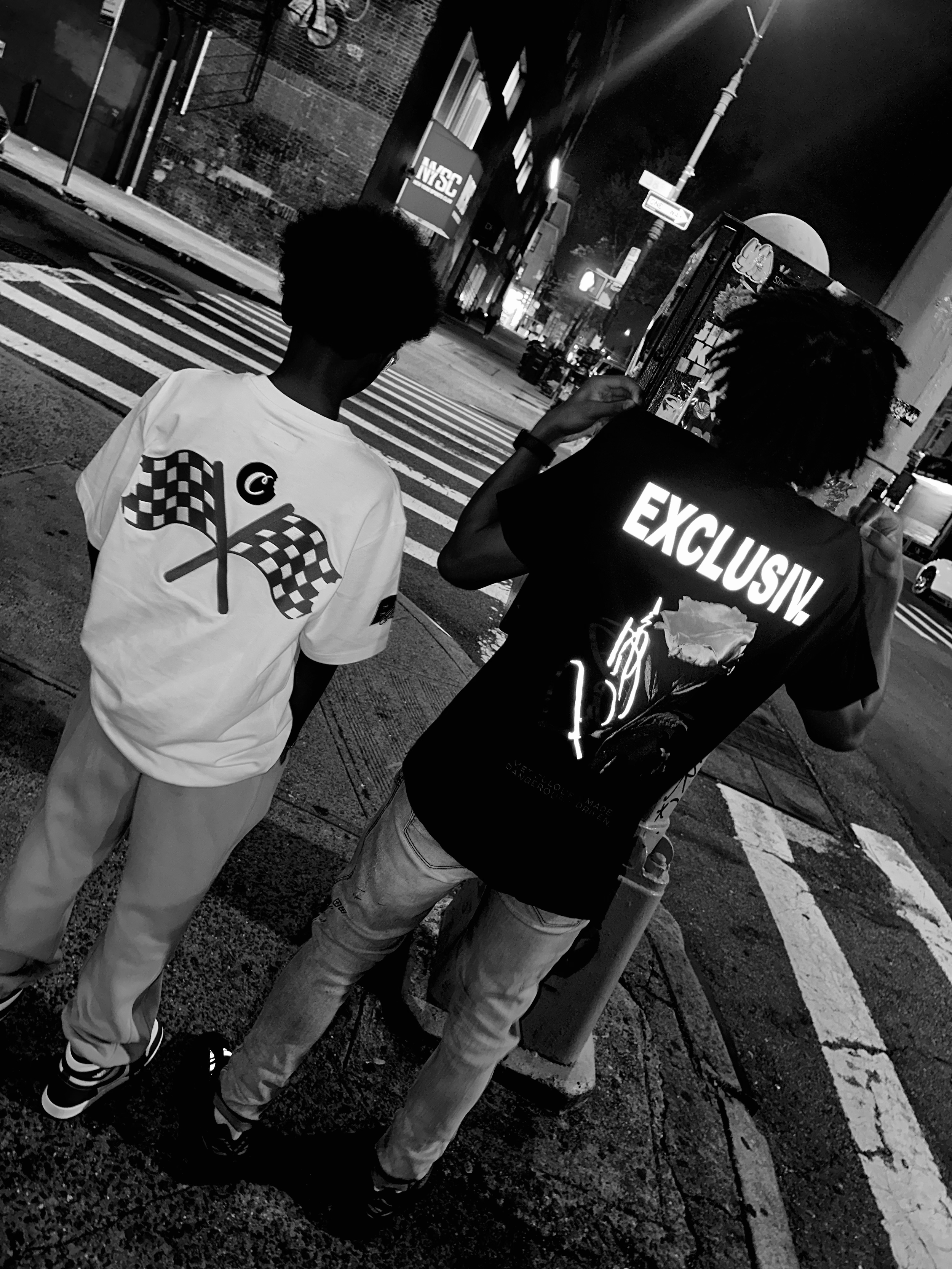 Two young men standing on a city street corner at night, one wearing a white T-shirt with checkered flags, the other wearing a black T-shirt with illuminated text reading 'EXCLUSIV' and graphics, illuminated city buildings and streetlights in the bac