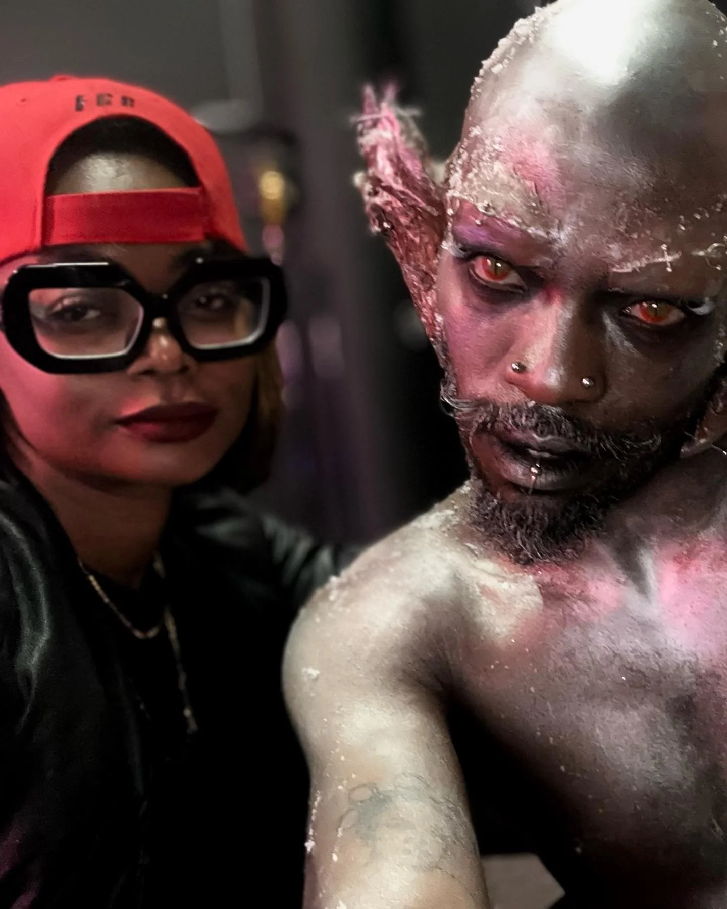 A woman wearing a red cap backwards, large black glasses, and a leather jacket taking a selfie with a person in elaborate makeup resembling a fantasy creature with metallic silver skin, pointed ears, red eyes, and facial piercings.