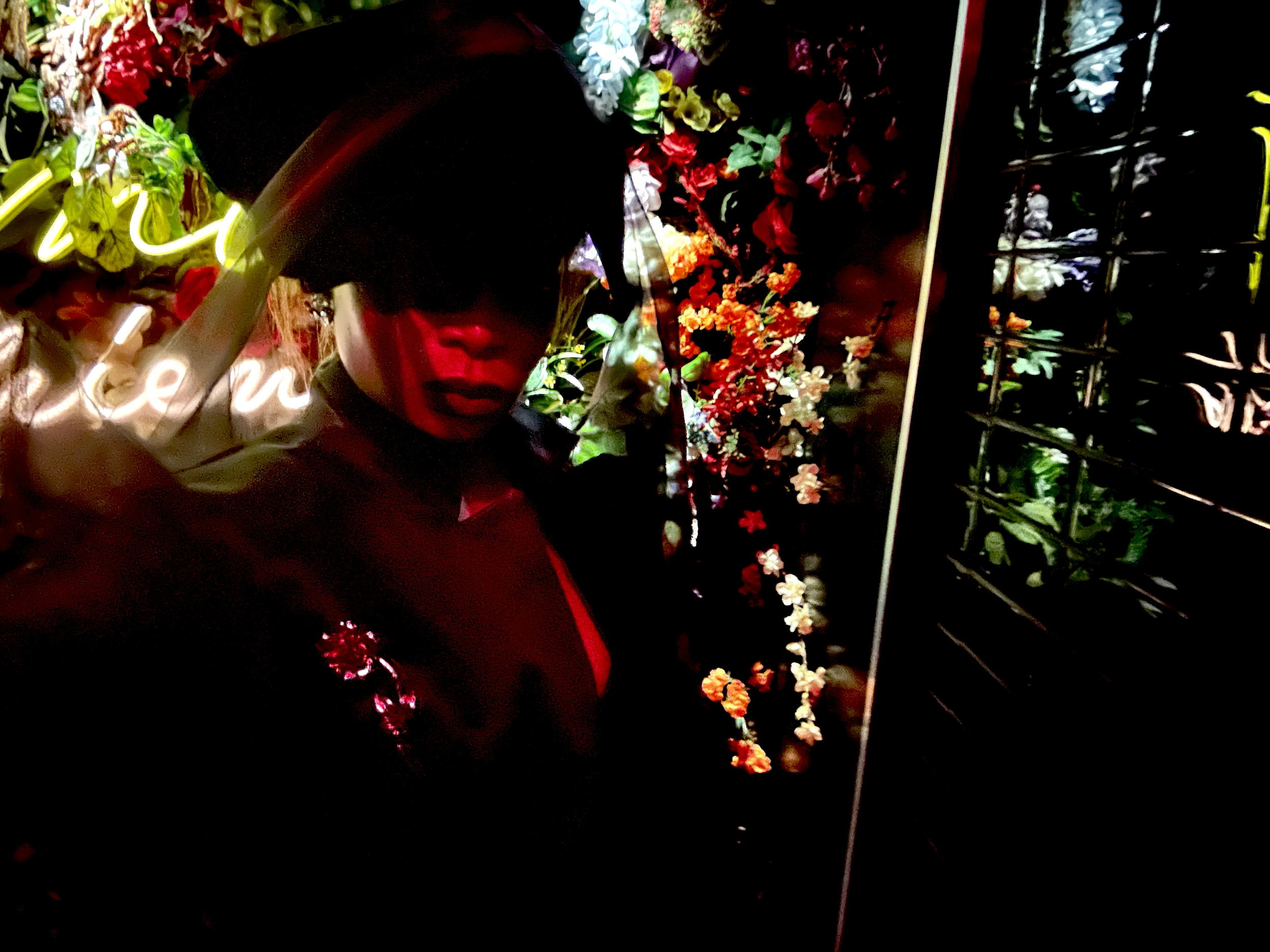 A person standing in front of a wall of colorful flowers and illuminated neon signs at night.