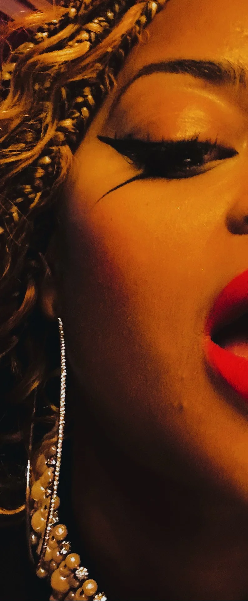 Close-up of a woman’s face showing makeup and jewelry, with curly hair on the left, bold eyeliner, and red lipstick.