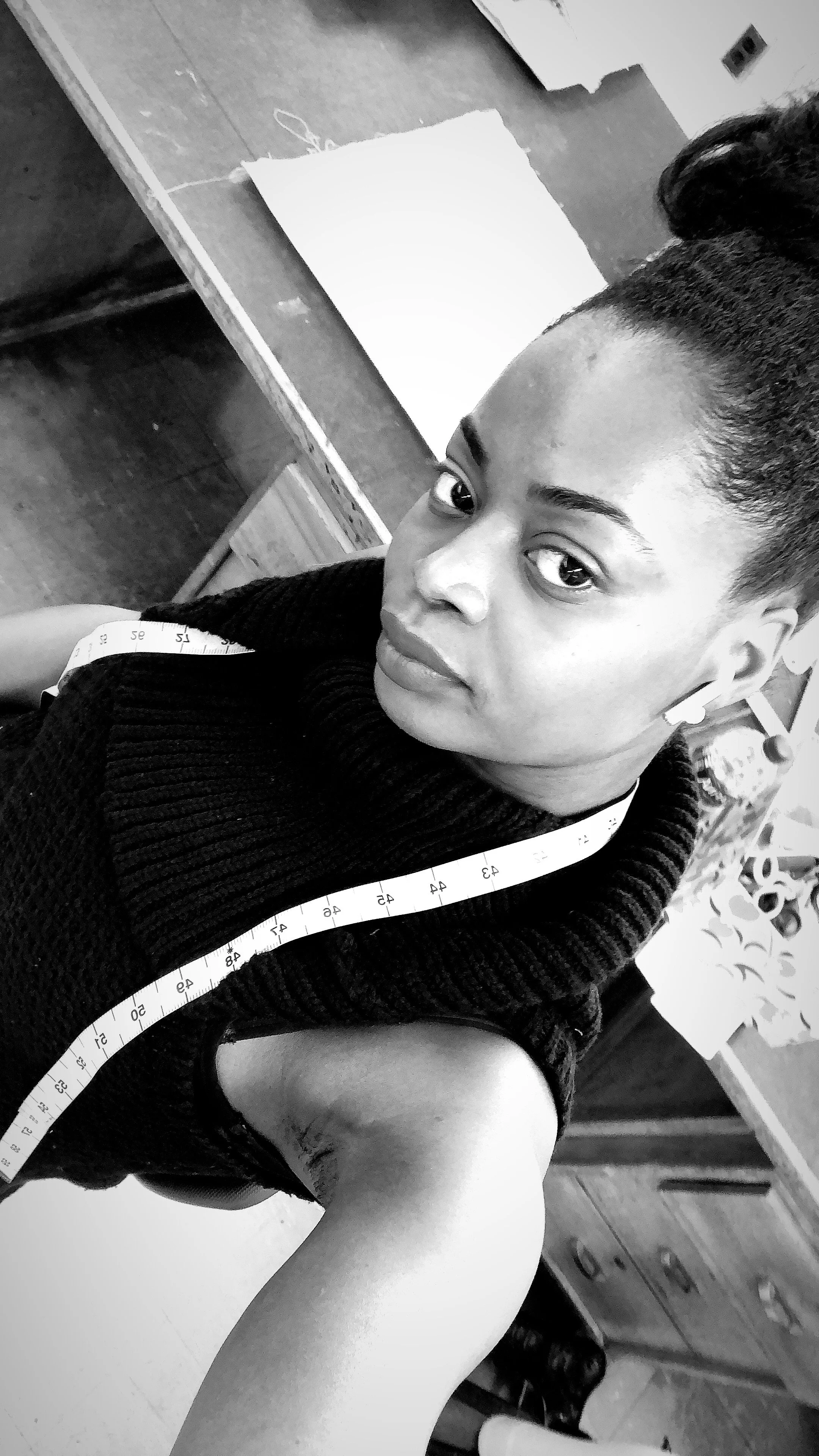 A woman taking a selfie in black and white, wearing a sleeveless top with a measuring tape around her neck, in a room with a wooden desk and papers.