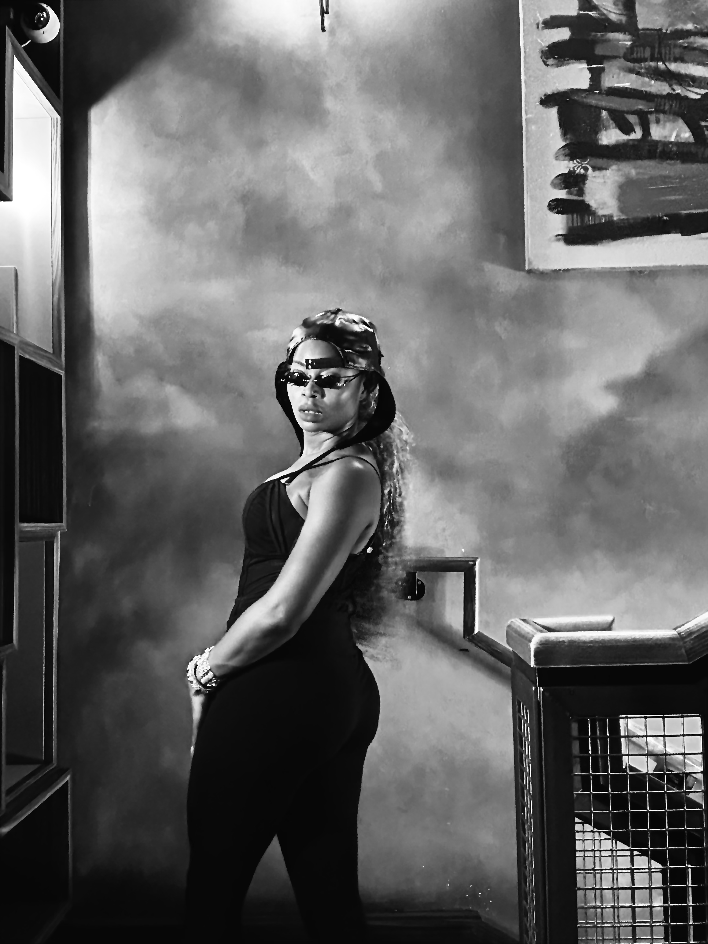 A woman with curly hair wearing sunglasses, a cap backwards, a black tank top, and black pants, standing indoors near a staircase with an open hand railing. There is a painting on the wall behind her and a black and white color scheme.
