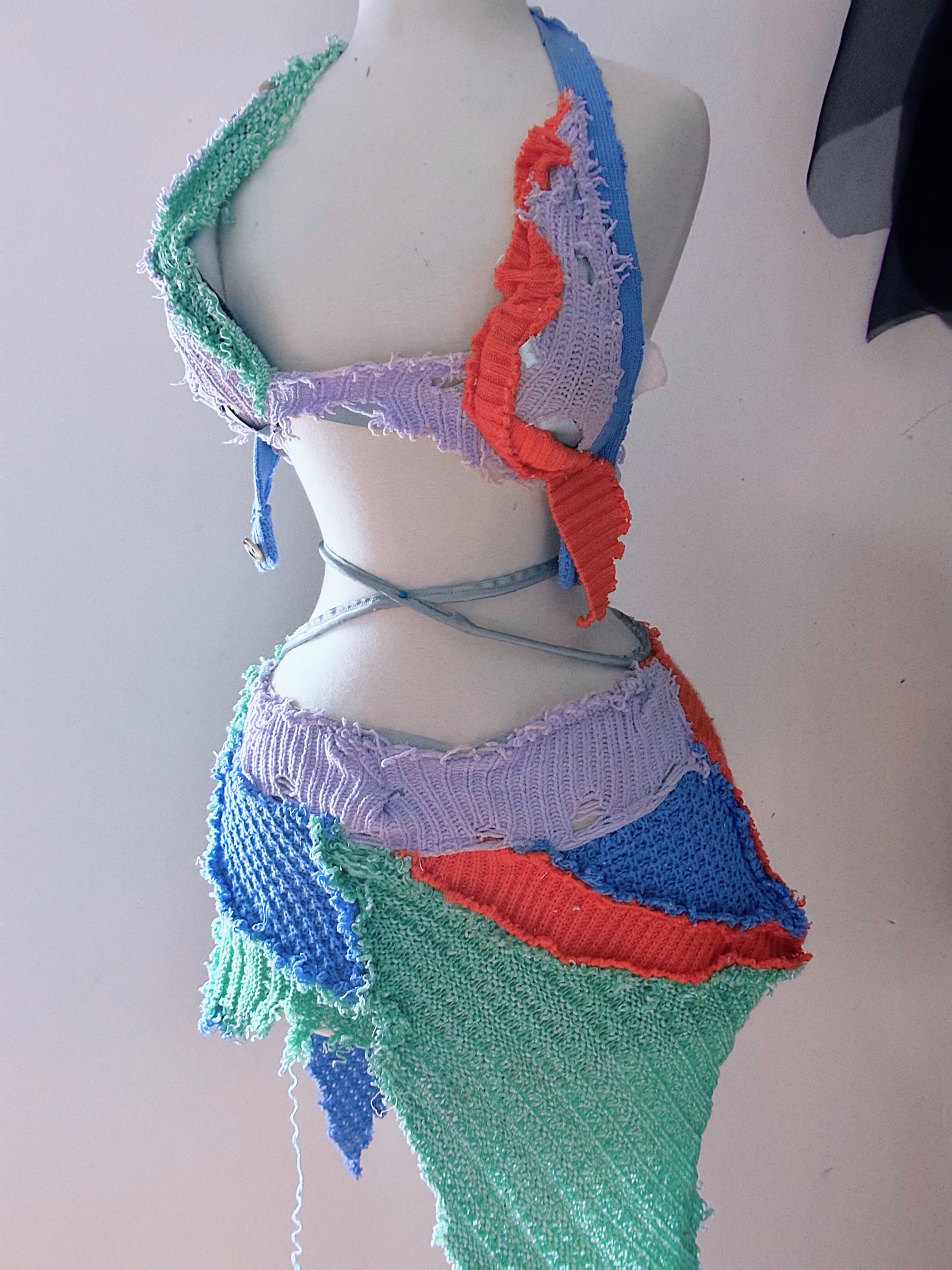 A mannequin dressed in a colorful, patchwork crochet outfit with different textured yarns and colors, resembling a skirt and top.