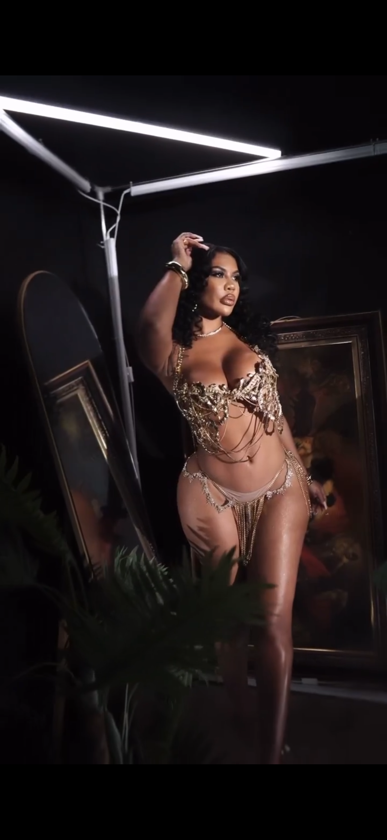 A woman in a gold and silver jewelry bikini standing in front of a mirror with a dark background and LED ceiling lights.
