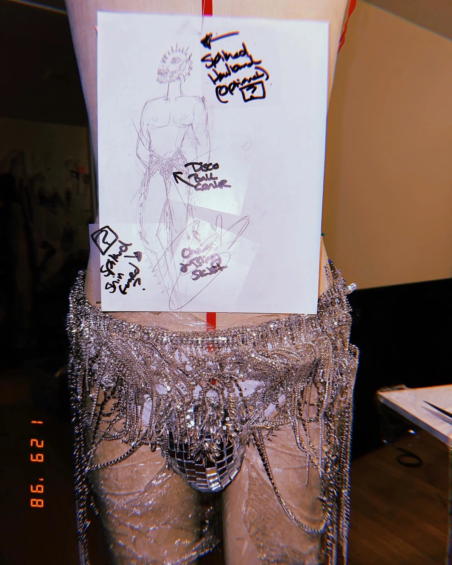 Sketch of a male figure wearing a body chain, notes indicating a 'disco ball center' and 'sand house,' with chains and jewelry draped over a mannequin dressed in rhinestone-embellished pants.