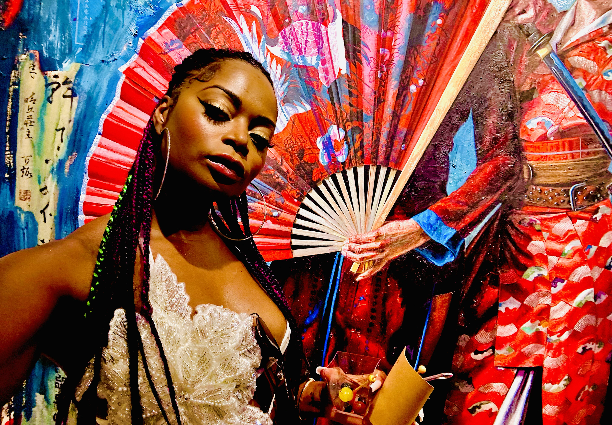 A woman with braided hair, wearing a white lace top and large hoop earrings, posing next to a colorful Japanese-style painted fan and an artwork of a person in red traditional clothing holding a fan.