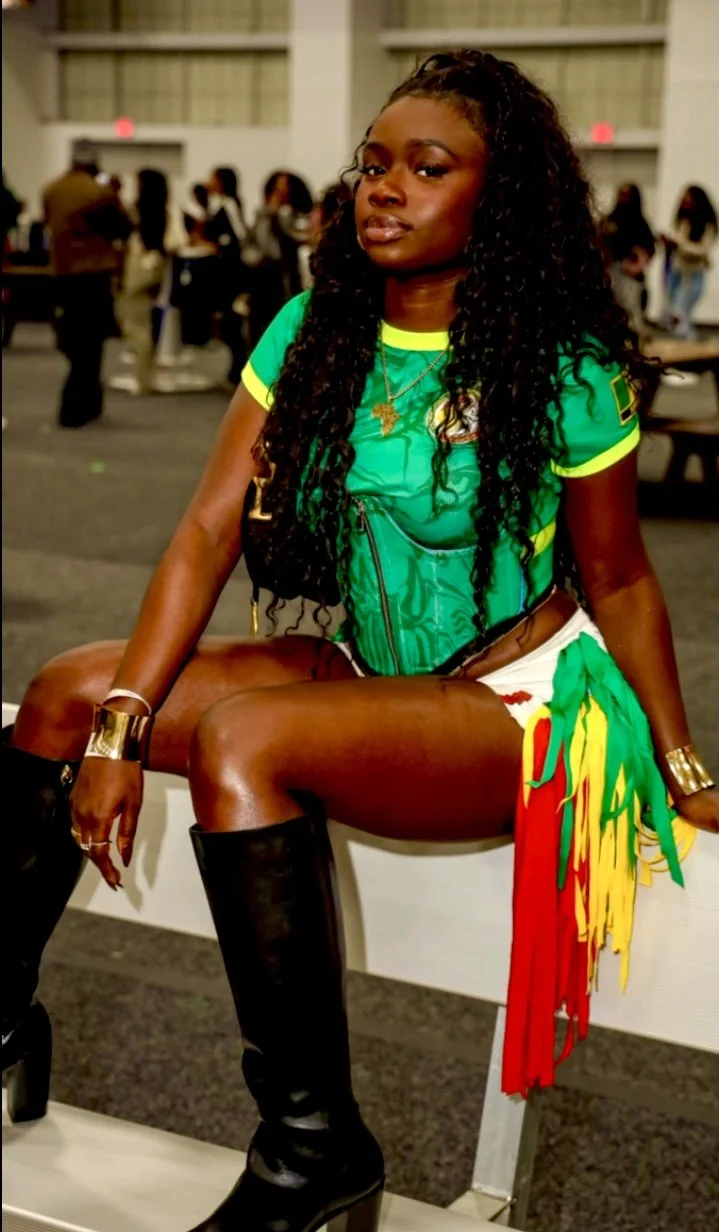 A woman with long curly hair, wearing a vibrant green sports jersey, thigh-high black boots, and colorful accessories, sitting on a table at an indoor event, with blurred people in the background.