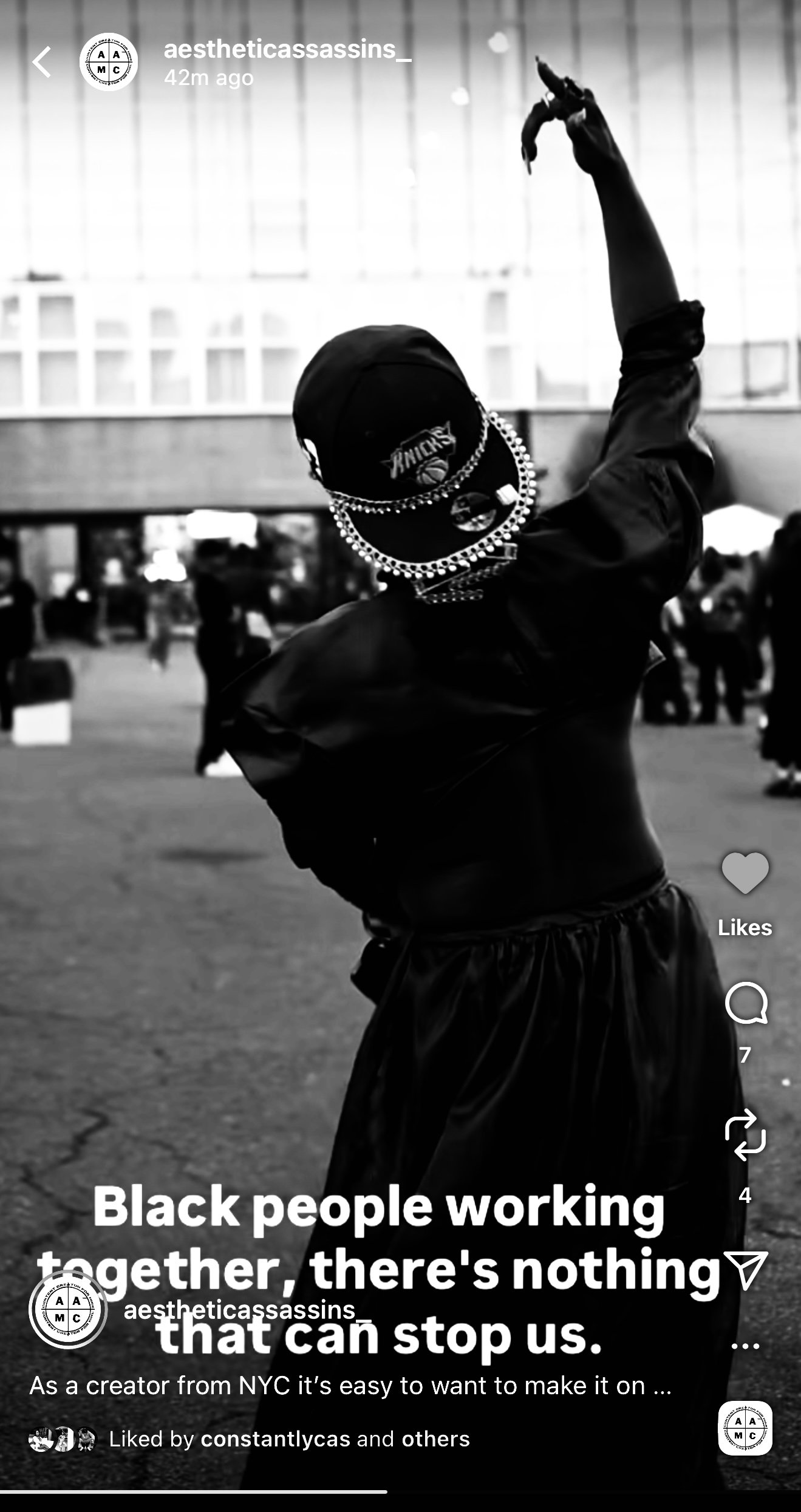 A person wearing a New York Knicks cap and jewelry, with their face hidden, standing outdoors in a crowd. The photo is in black and white.