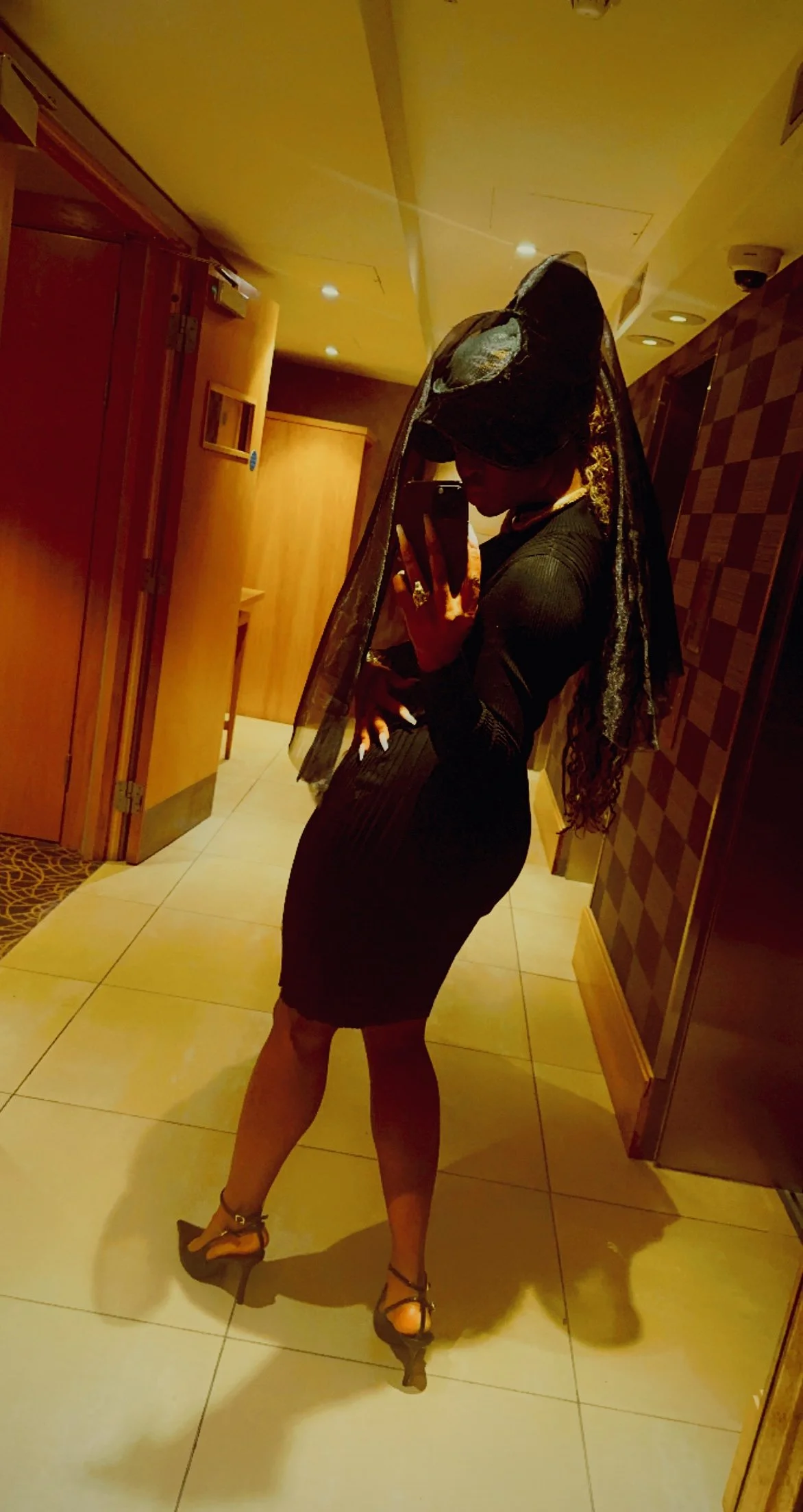 A woman taking a mirror selfie in a dimly lit hallway, wearing a black dress, high heels, and a lace veil over her head.