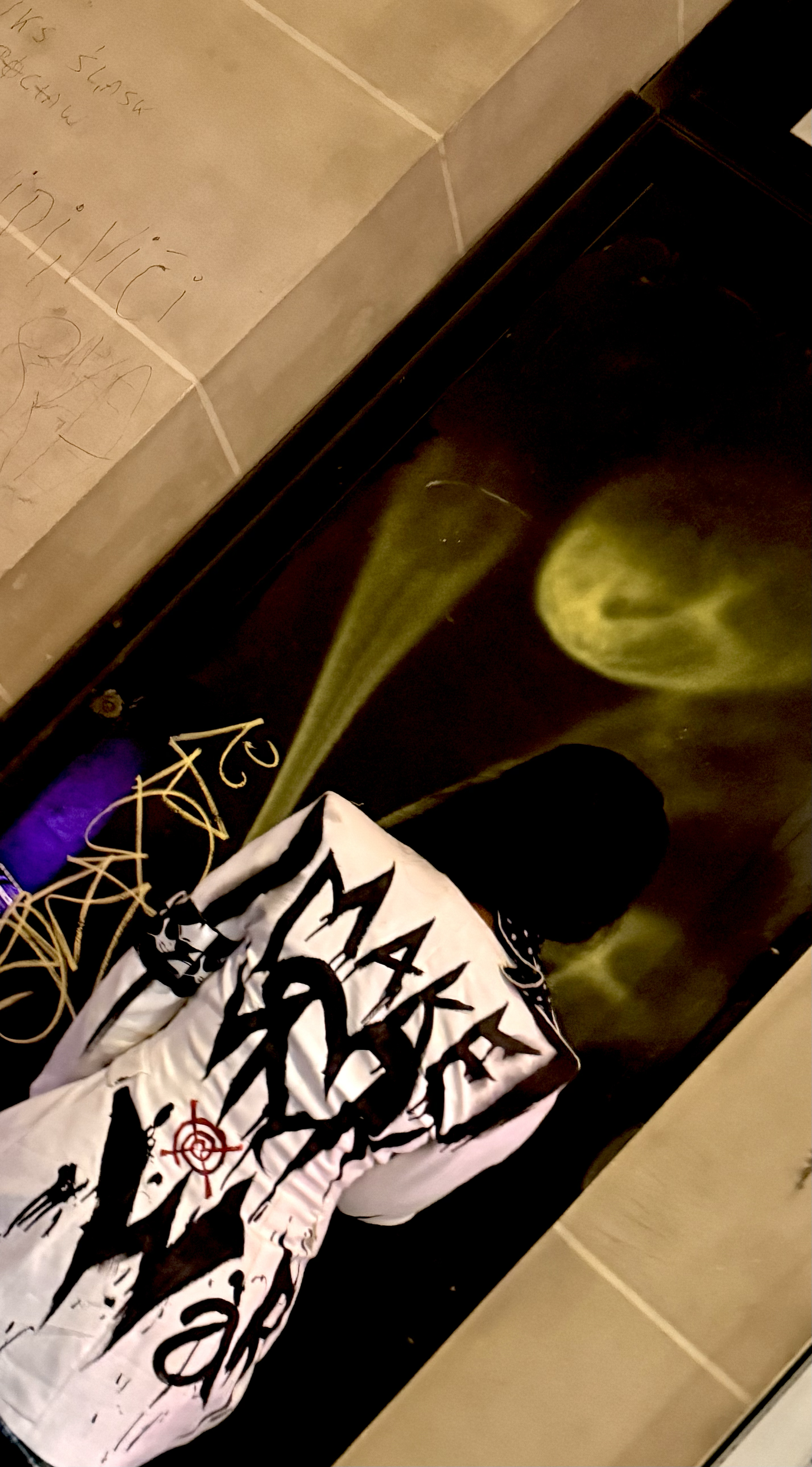 A person wearing a white jacket with black handwritten-style graphics and text, standing on a black floor with green and yellow spore-like designs, near tiled walls with some scribbles and notes.