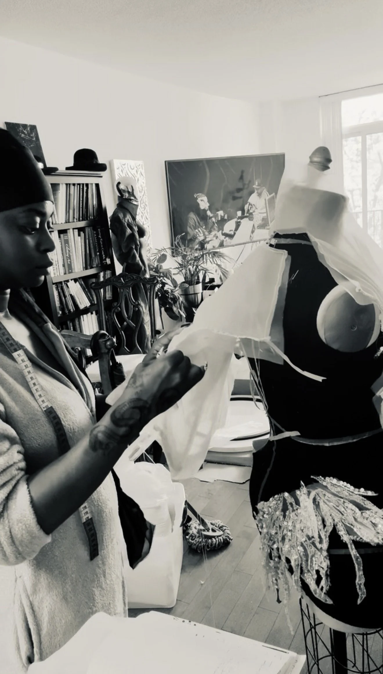 A woman sewing fabric on a dress form in a well-lit room with bookshelf, plants, and artwork in the background.
