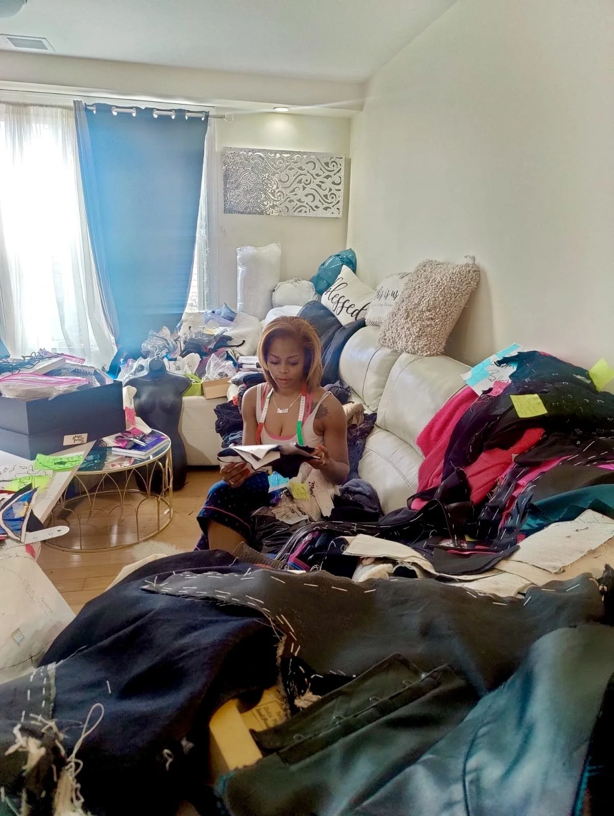 A woman sitting on a cluttered sofa reading a book in a living room with piles of clothes and belongings surrounding her.