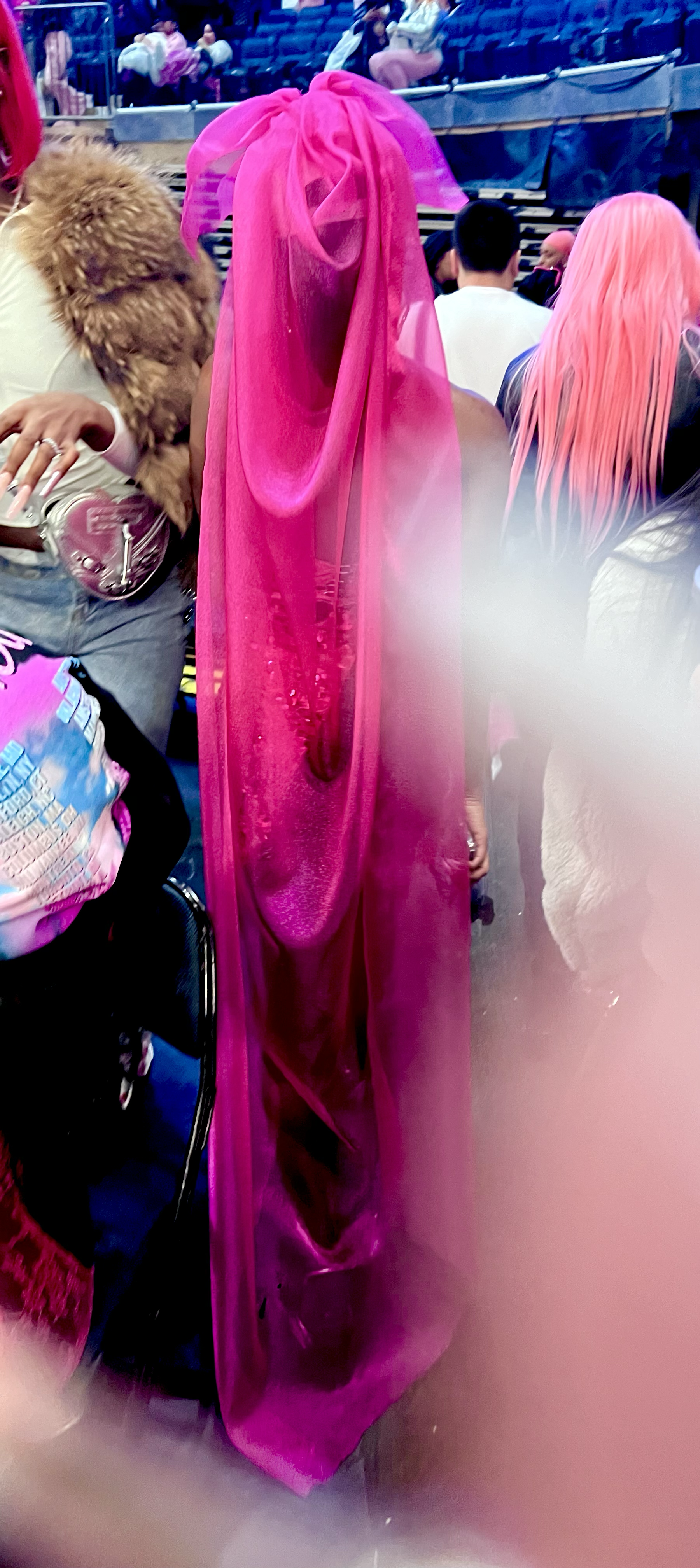 Person dressed in a bright pink, flowing, sheer costume resembling a jellyfish, standing among other people at an indoor event or convention.