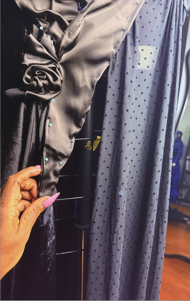 Close-up of a hand with pink nails holding a piece of black satin fabric with pinned blue and red push pins, next to a sheer gray fabric curtain with black polka dots.