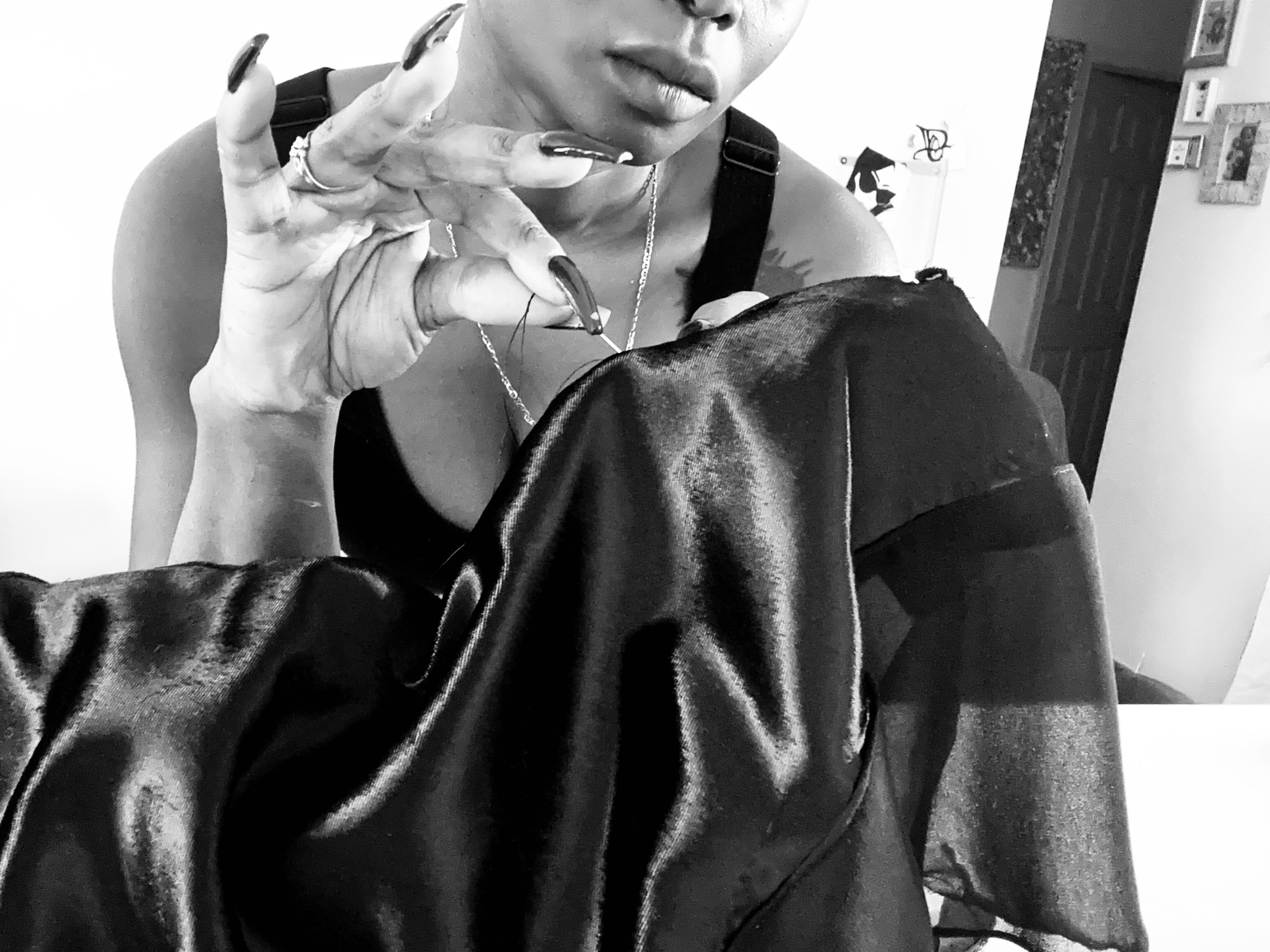 A woman in a black top is holding a sewing needle, working on a black shiny fabric, possibly leather or vinyl.