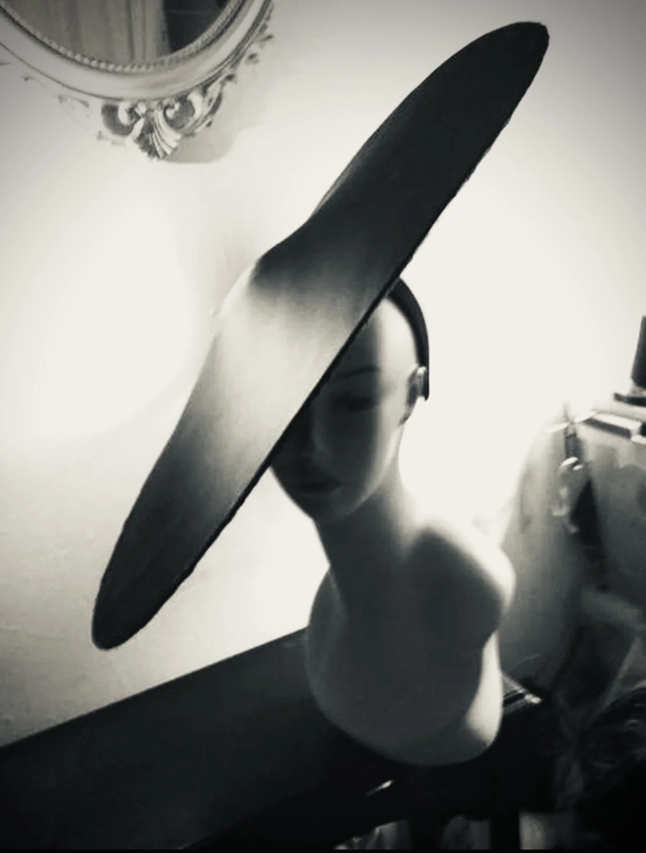 Black and white photo of a woman partially hidden behind a large round mirror, with her face visible through the mirror's reflection, standing next to a dresser and a lamp.