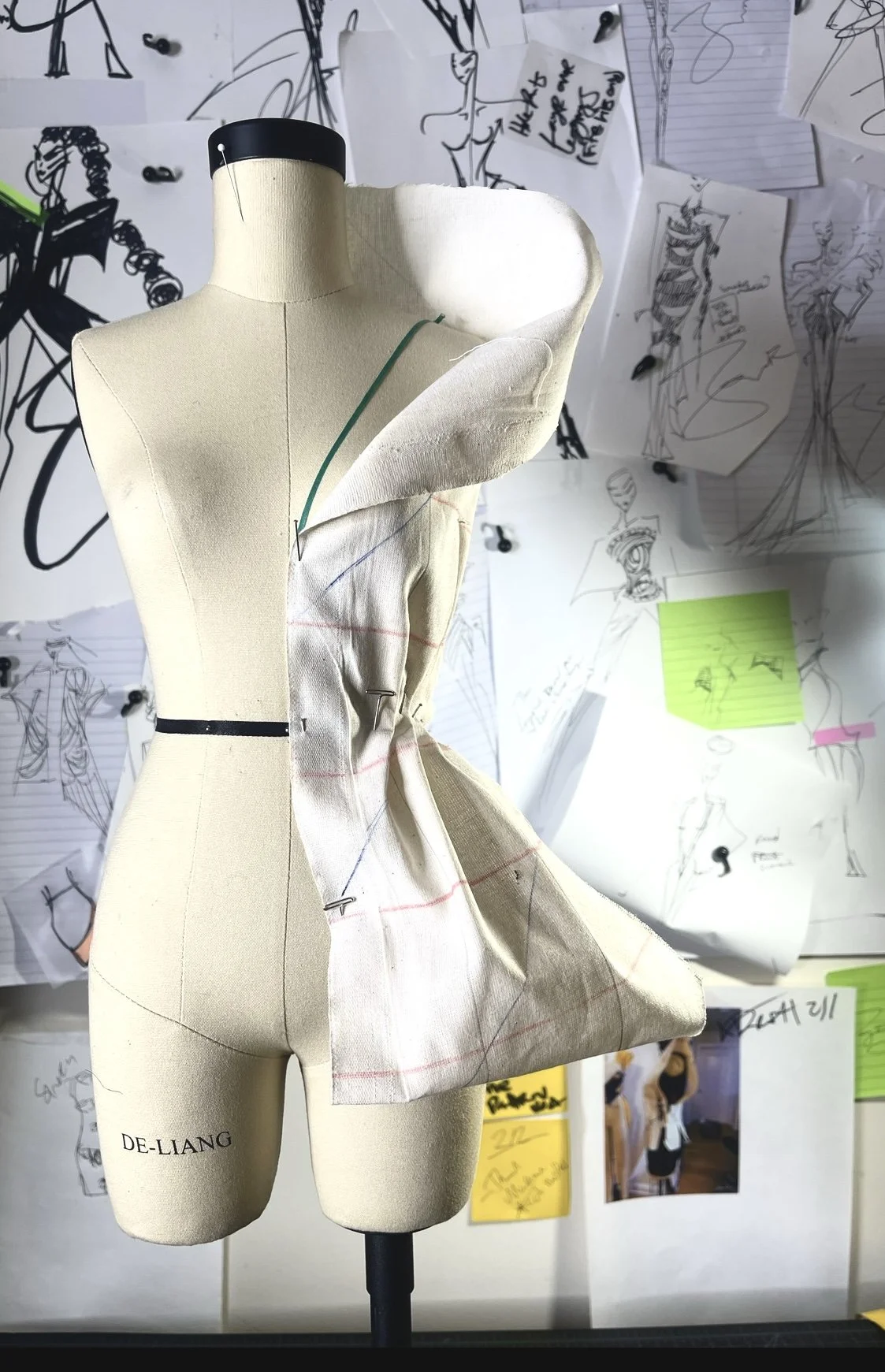 A dress form labeled 'De-Liang' with fabric and pattern markings, surrounded by sketches and notes pinned on a wall.