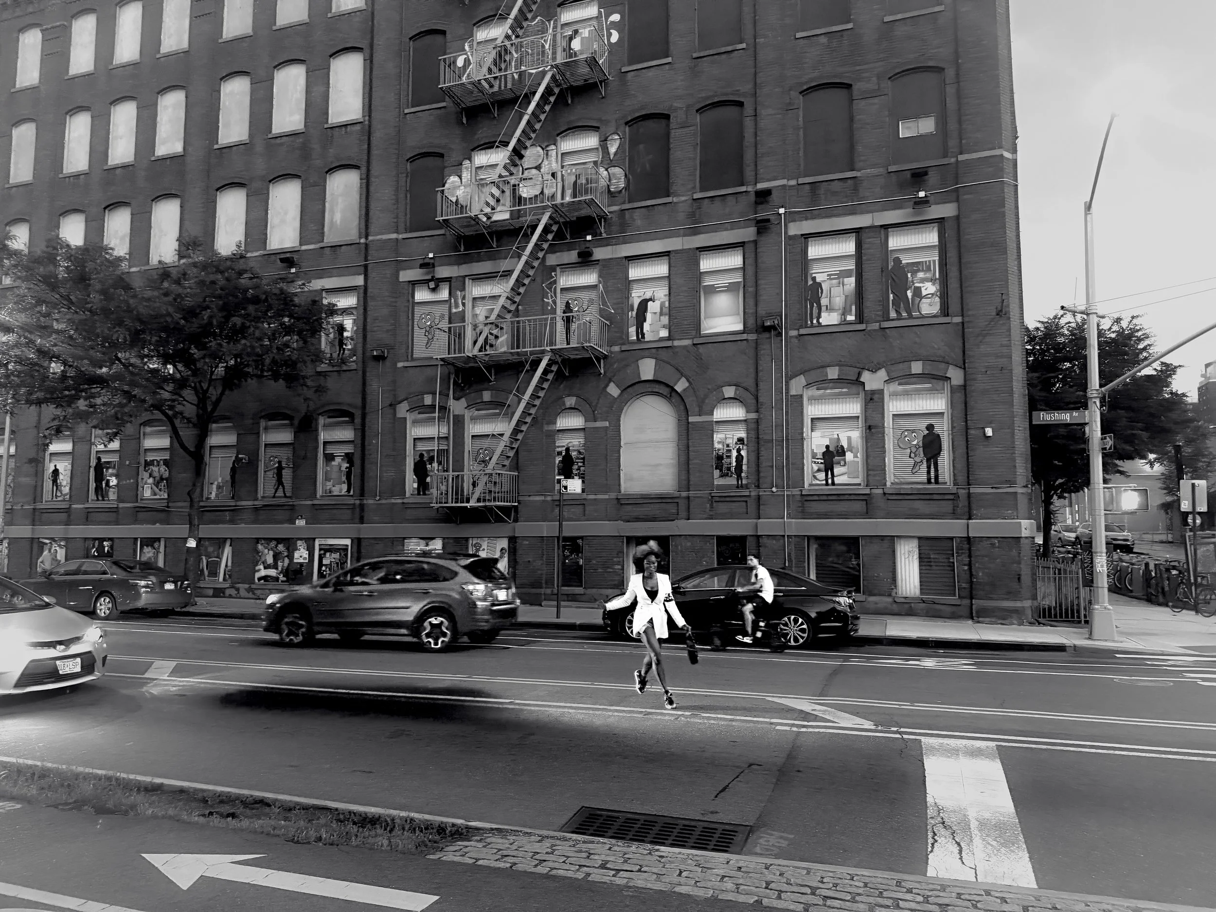 A city street scene in black and white, showing a multi-story brick building with fire escapes, pedestrians on the sidewalk and crossing the street, and cars passing by. There are people visible through the windows of the building.