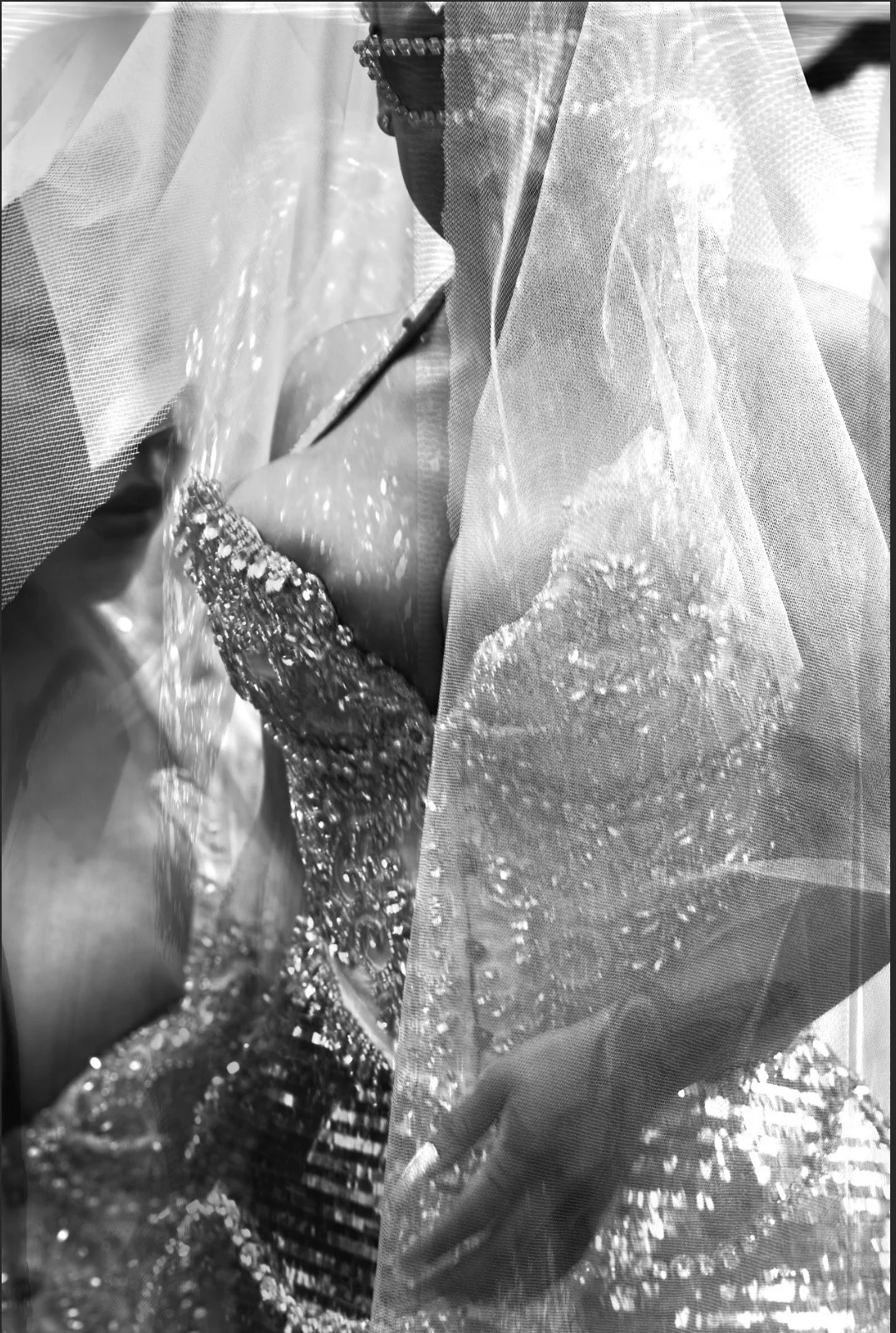 Close-up of a woman wearing an embellished beaded wedding dress with a lace veil covering her face, seen through a sheer fabric.