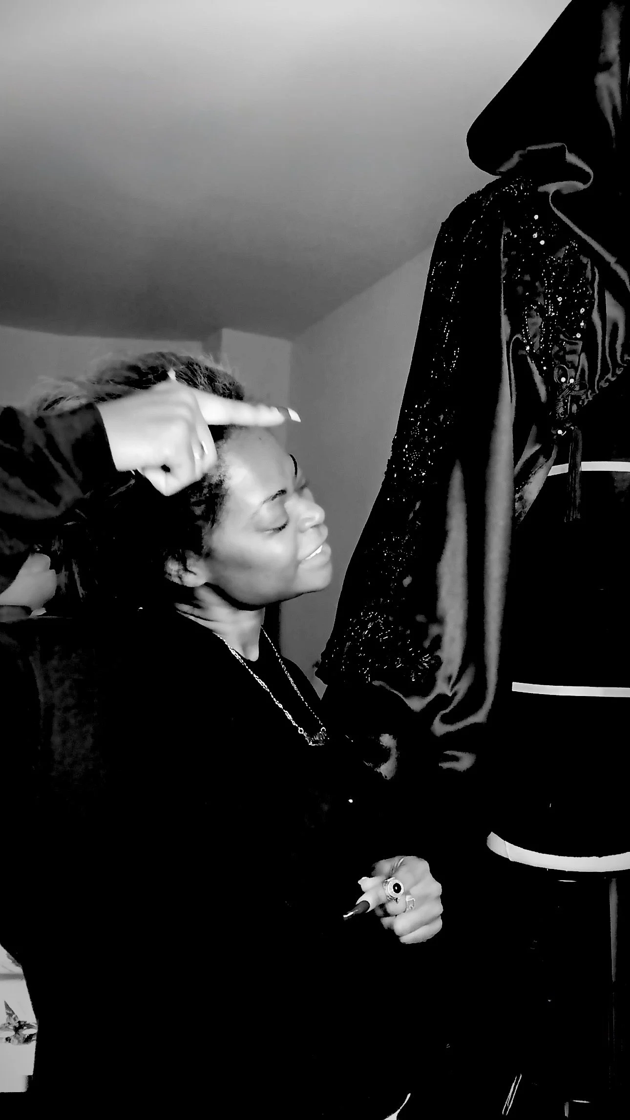 A woman smiling with eyes closed, sitting on a chair, while another person applies makeup or face paint on her forehead. The scene is in black and white.