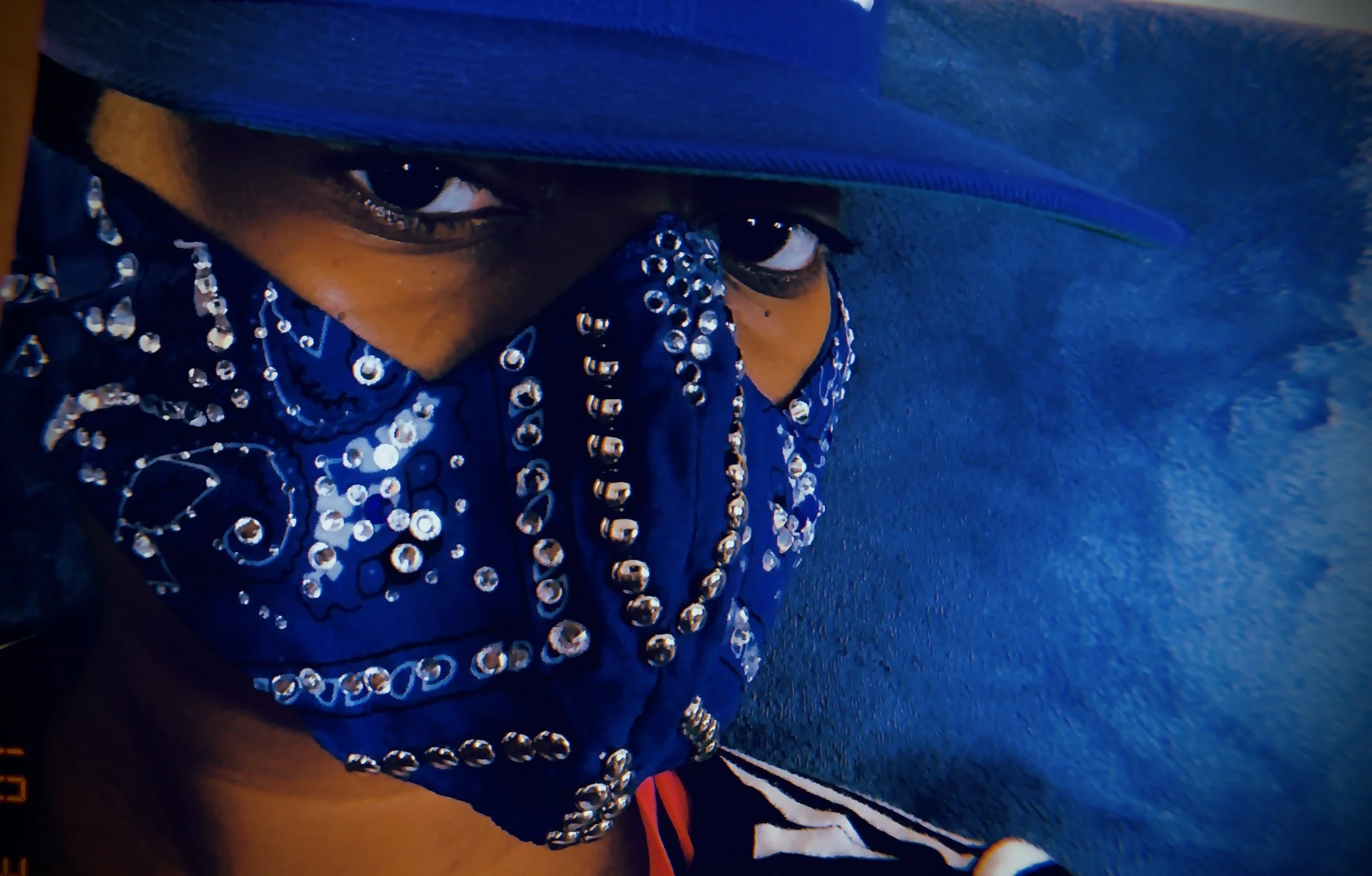 Close-up of a person wearing a blue bandana with silver embellishments covering their mouth, and a blue cap, with their eyes visible, on a blue background.
