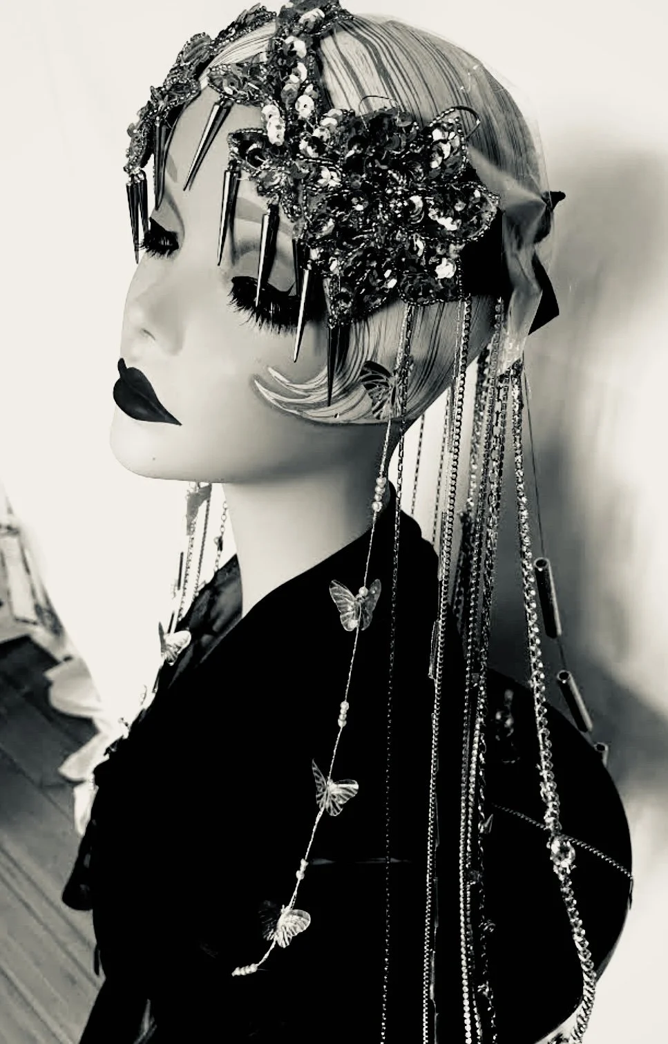 Black and white photo of a mannequin head wearing a detailed headpiece with sequins and chains, with long butterfly-shaped earrings and dark lipstick.