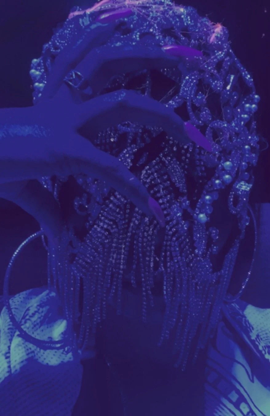 Close-up of a person with long hair, wearing a black hooded jacket with reflective dots, in a dark setting with blue and purple neon lighting.