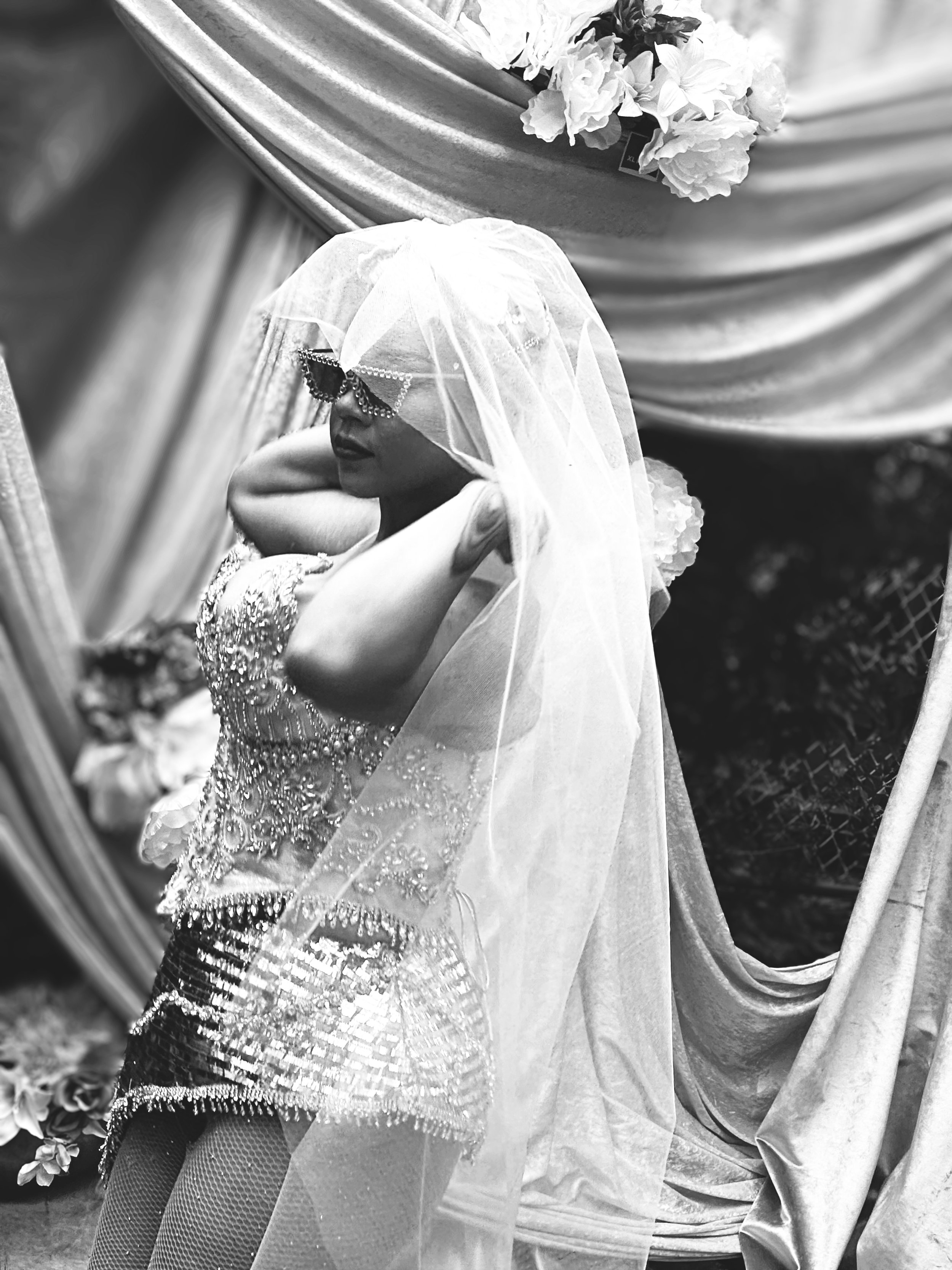 A woman in an ornate, beaded dress and fishnet stockings, wearing sunglasses and a veil, poses with her arms behind her head, standing in front of a draped fabric backdrop with flowers.