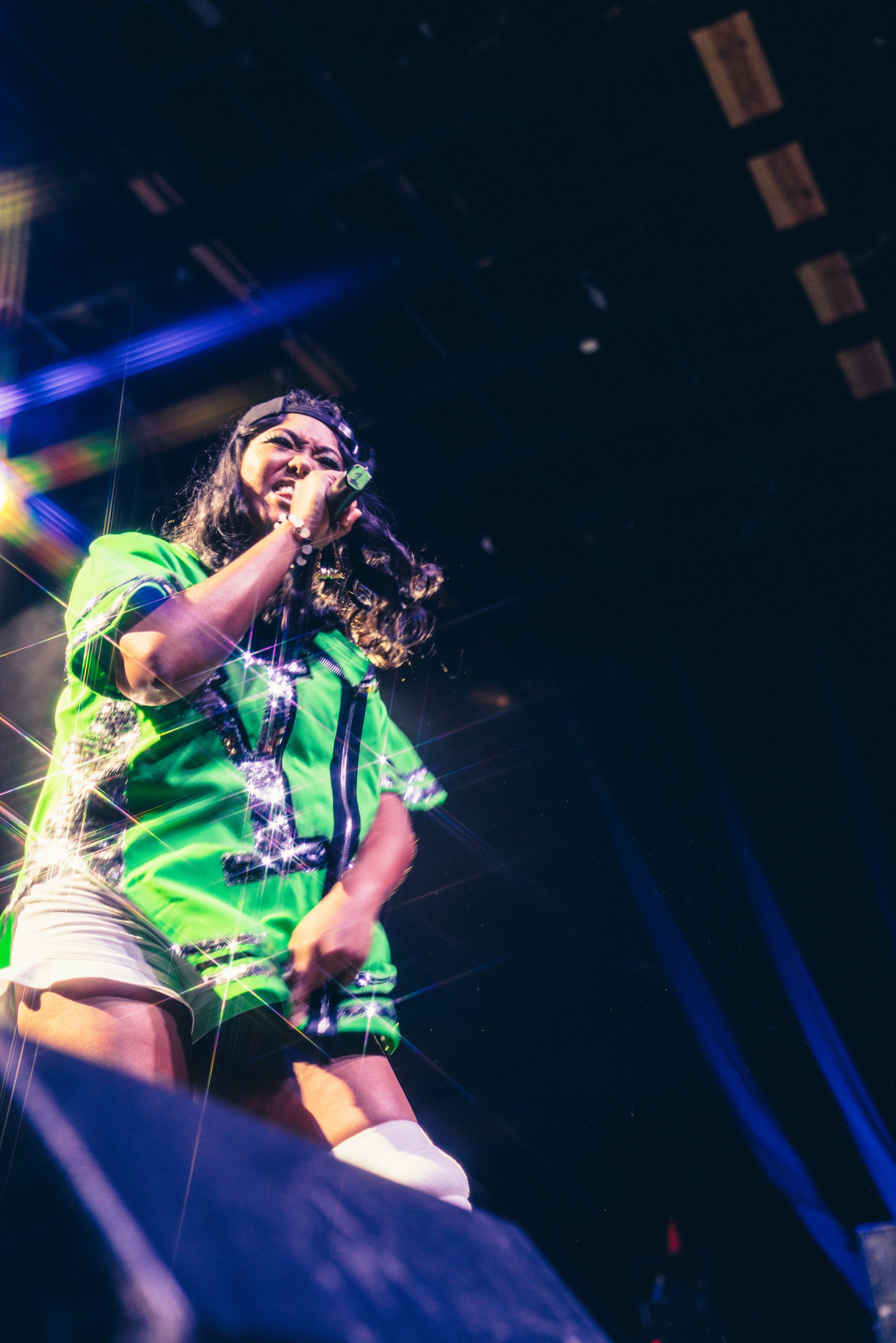 A woman performing on stage, holding a microphone, wearing a bright green jacket, shorts, and knee-high socks, with colorful stage lighting and effects.