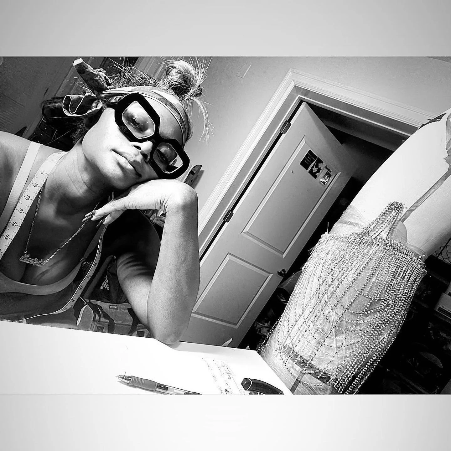 A woman wearing safety goggles and a lightweight top, sitting at a table with a measuring tape around her neck, resting her chin on her hand. In the background, there is a dress form adorned with numerous strings of beads or pearls, in a room with a 