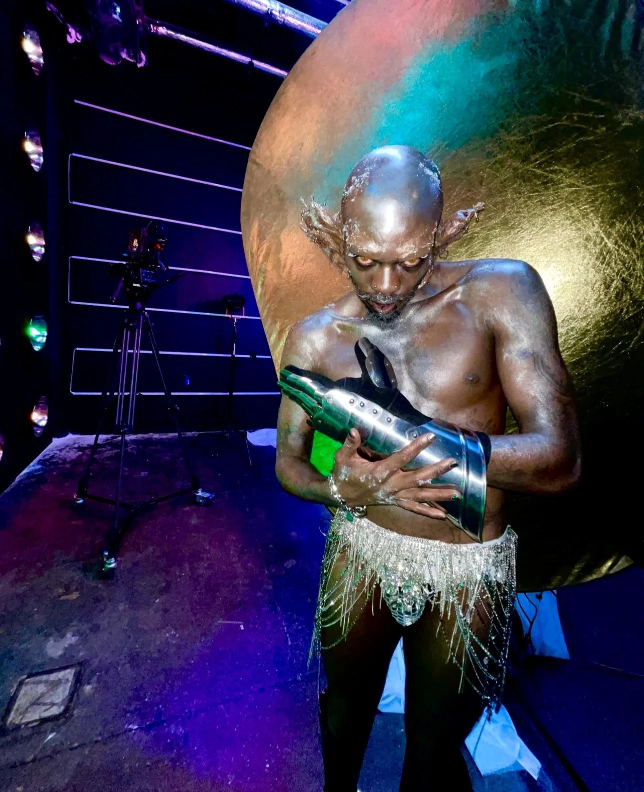 A shirtless person with a shaved head, eccentric makeup, and earrings holding a shiny prop, standing in front of a colorful, textured backdrop with stage lighting and equipment visible.