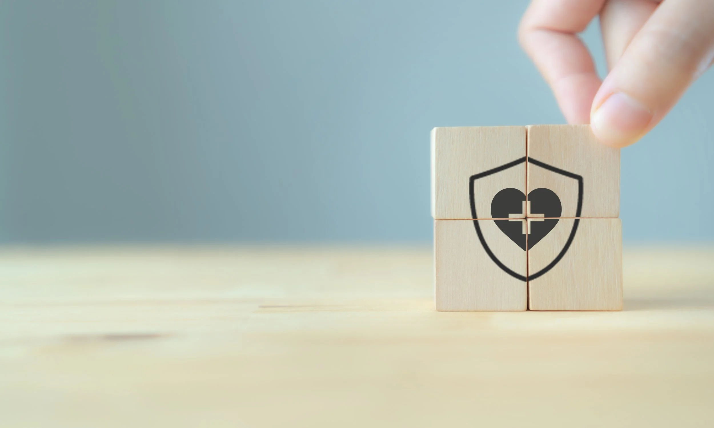 A hand placing wooden blocks with a health shield and broken heart symbol on a light wooden surface.