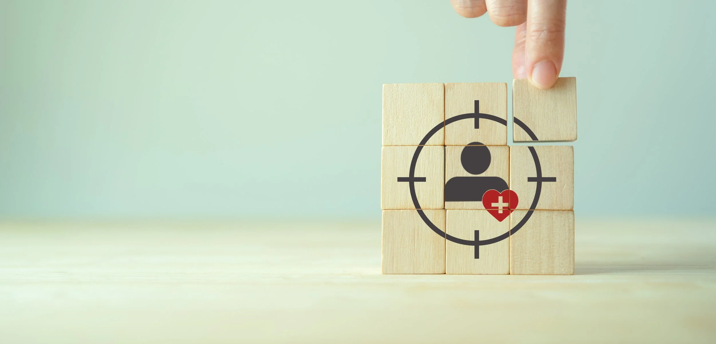 Stacked wooden blocks with a target symbol and a person icon, with a heart containing a medical cross on it, inside the target.