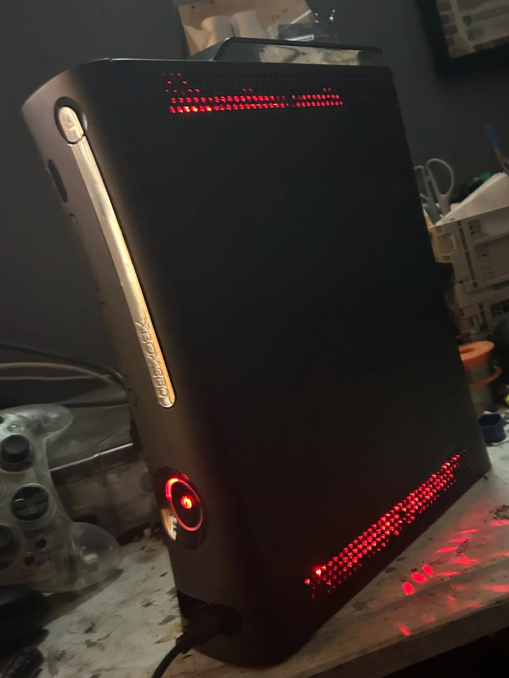 A black gaming console with red LED lights on a cluttered desk.