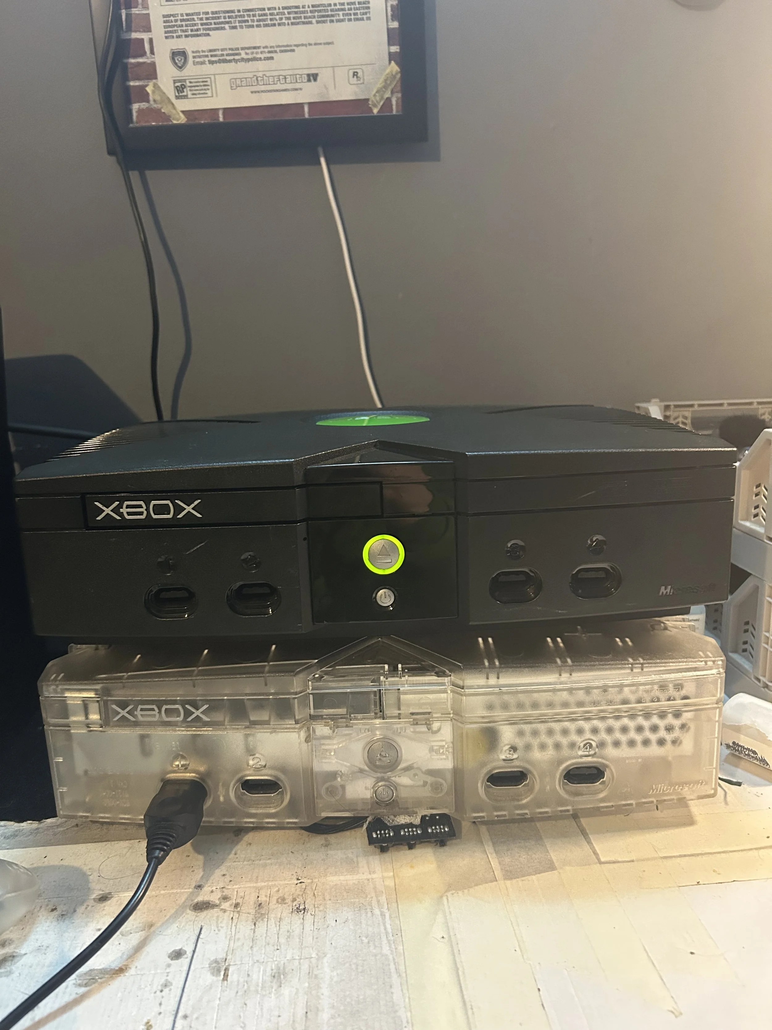 A black Xbox console stacked on top of a translucent Xbox console on a messy white wooden surface. A black power cord is plugged into the lower console. There is a framed poster with a partially visible notice and logos on the wall behind them.