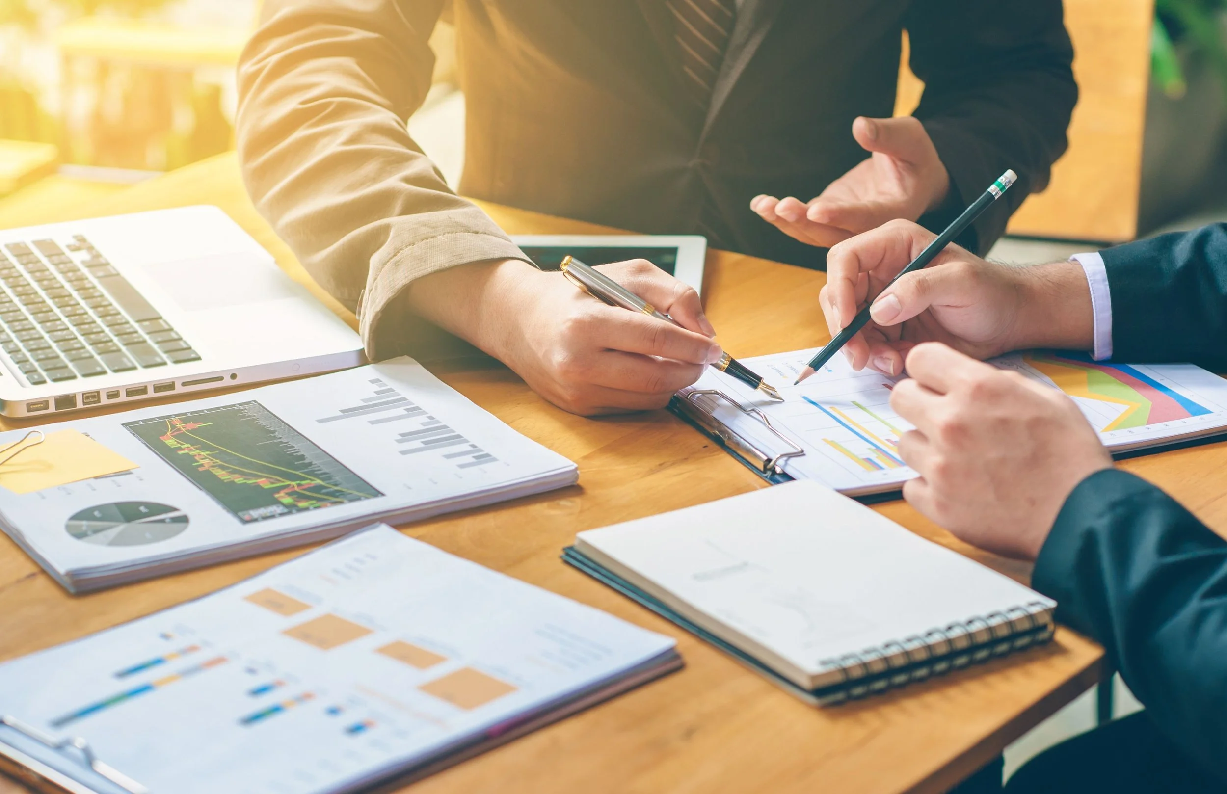Two individuals in business attire working on financial documents and charts at a wooden table with a laptop, notebooks, and printed graphs.