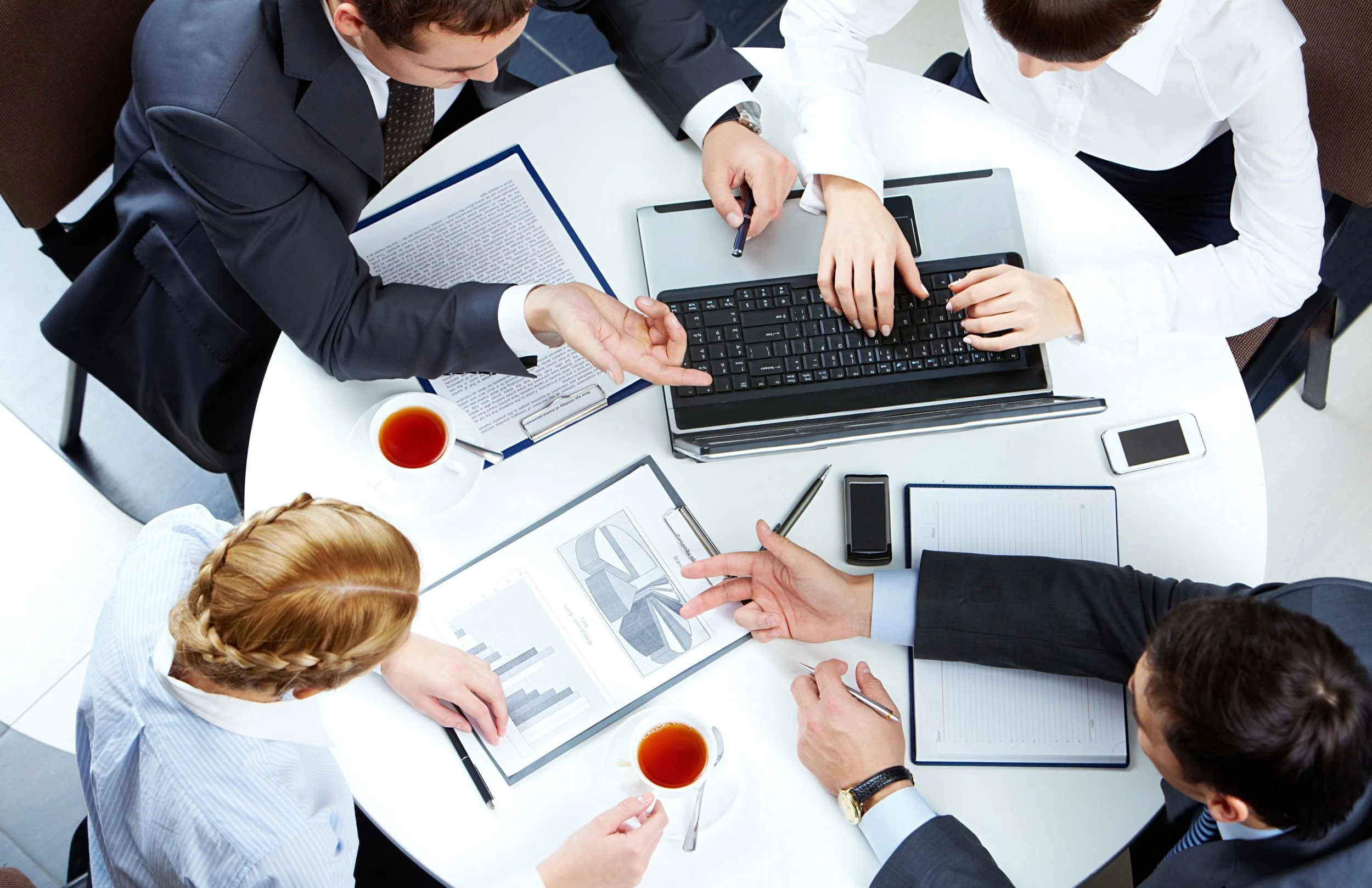 Top-down view of five professionals engaged in a business meeting around a white round table, discussing charts and documents, with laptops, smartphones, and cups of tea.