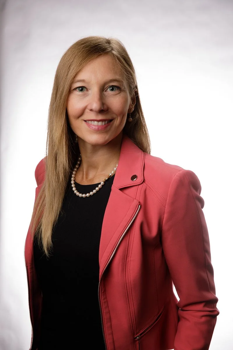 A woman with long blonde hair smiling, wearing a black top, a pink jacket, a pearl necklace, and earrings against a neutral background.