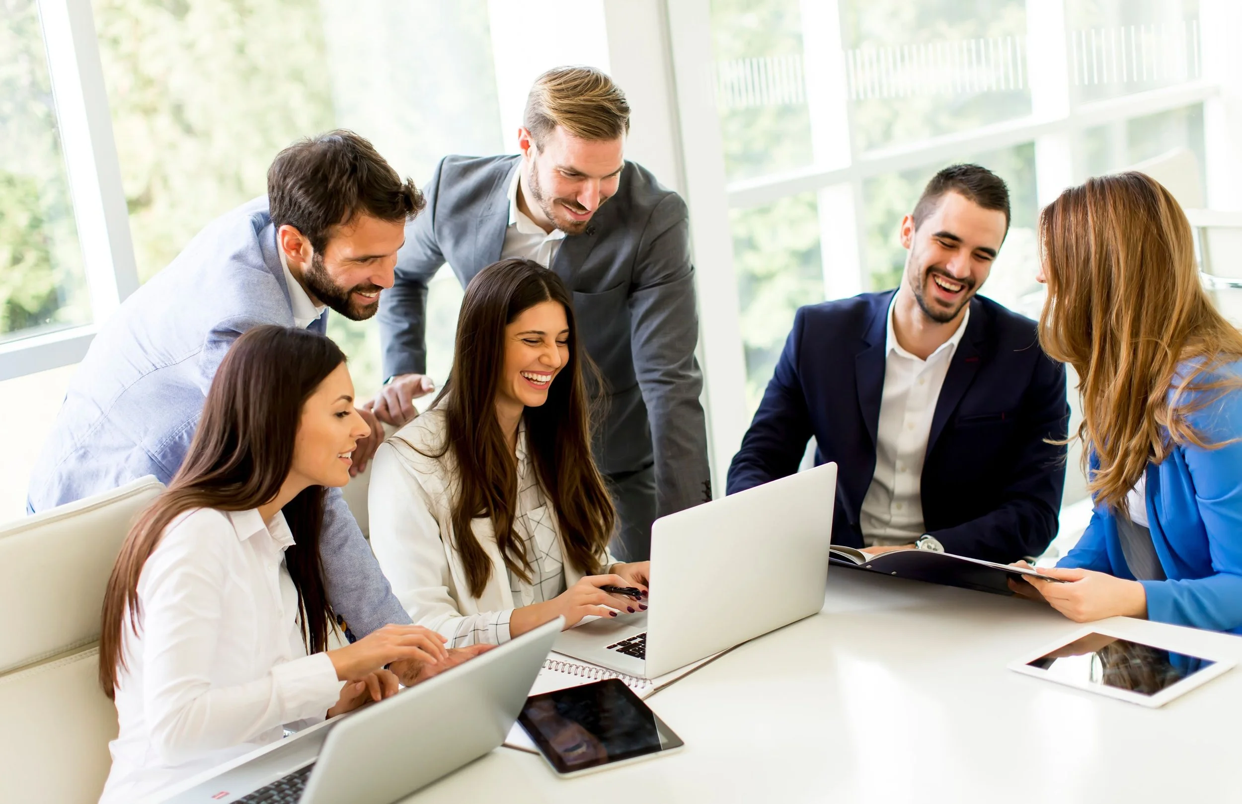 Group of six coworkers in a bright office, gathered around a table, smiling and looking at a laptop screen.