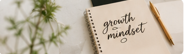 A spiral notebook with the words 'growth mindset' written on the page, a gold pen placed beside it, and a green plant blurry in the background.