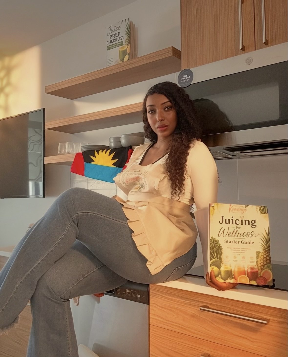 Woman sitting on kitchen counter next to a juicing wellness starter guidebook, in a modern kitchen with a flag of Antigua and Barbuda on a shelf behind her.