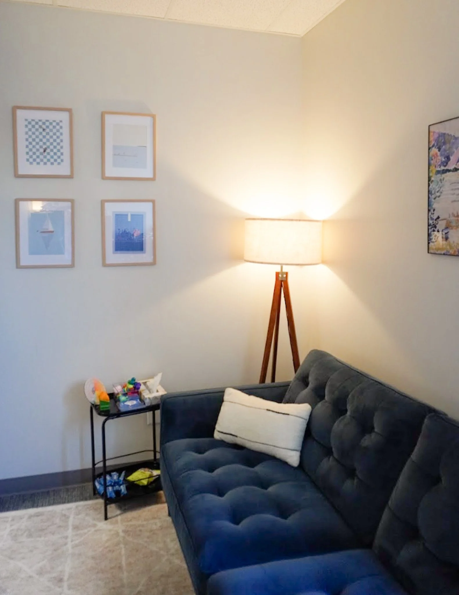 Cozy room with a blue sofa and white pillow, wall art in wooden frames, a floor lamp, and a side table with colorful toys, conveying a calm atmosphere.