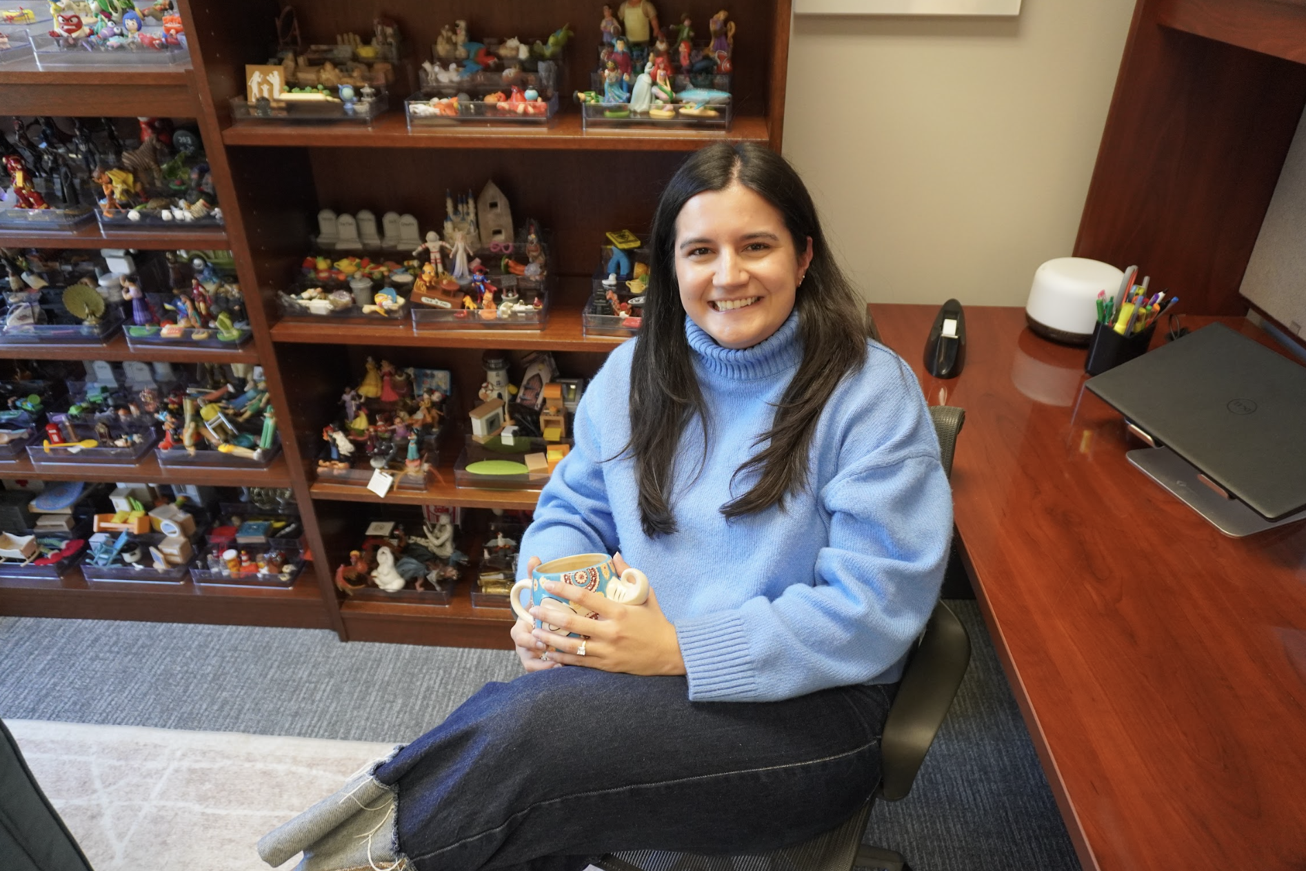 Smiling woman in a blue sweater sits on a chair, holding a mug. Behind her, shelves display colorful figurines.