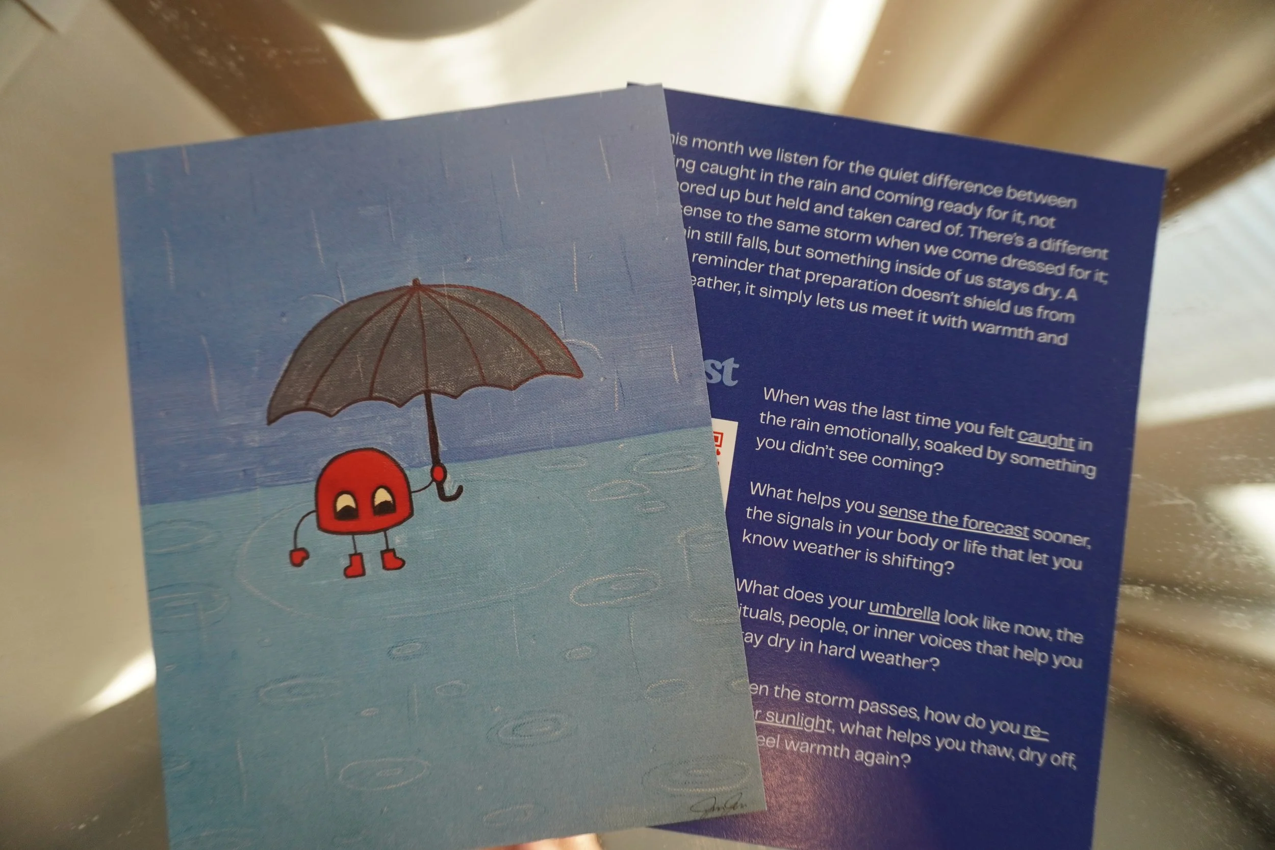 Two cards, one shows a cute red character with eyes, holding an umbrella on a blue background. The other card has text on it that is partially visible.