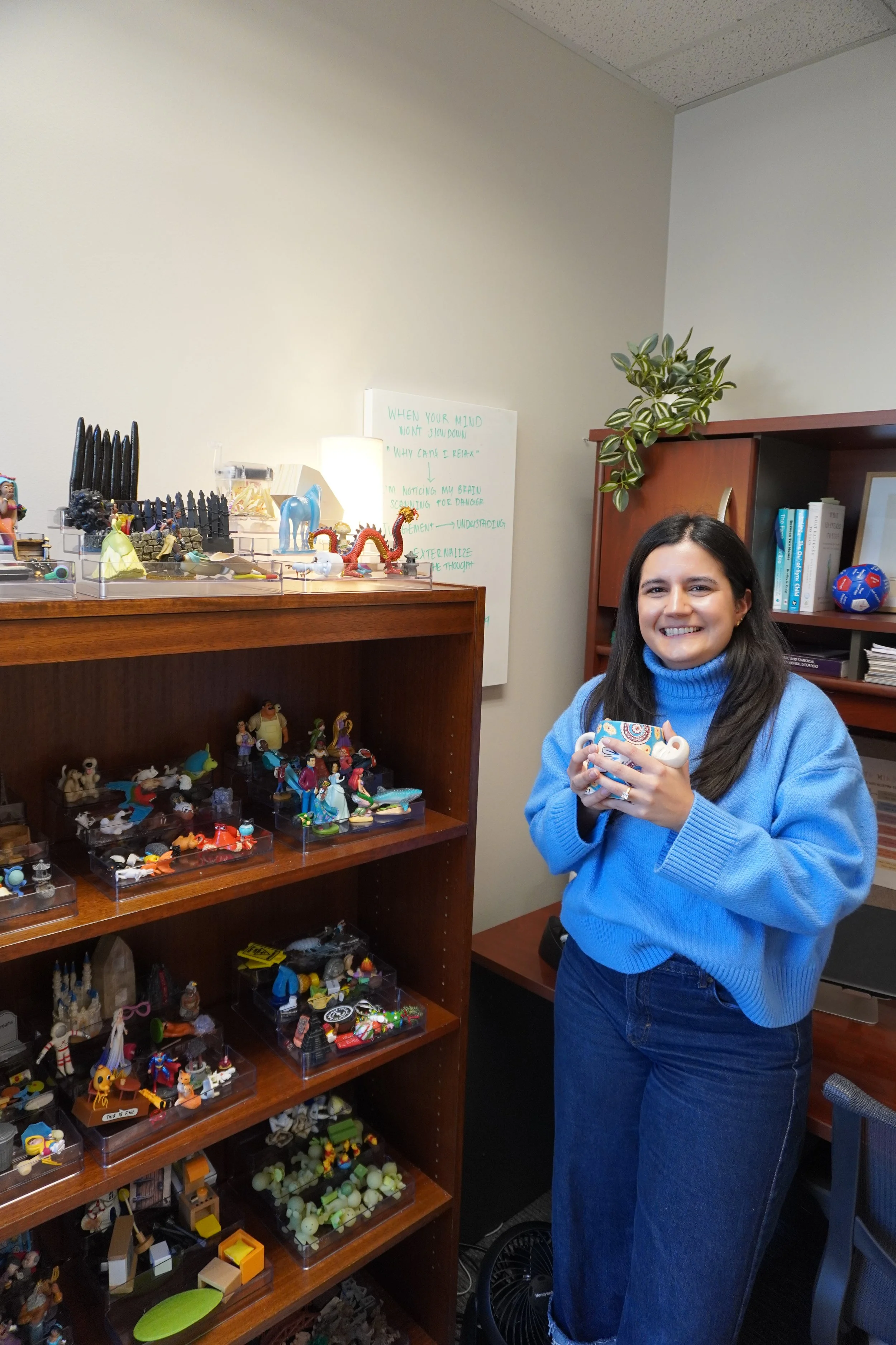 Woman in a blue sweater holding a colorful mug while standing in an office next to a wooden bookshelves filled with small figurines, books and a potted plant.