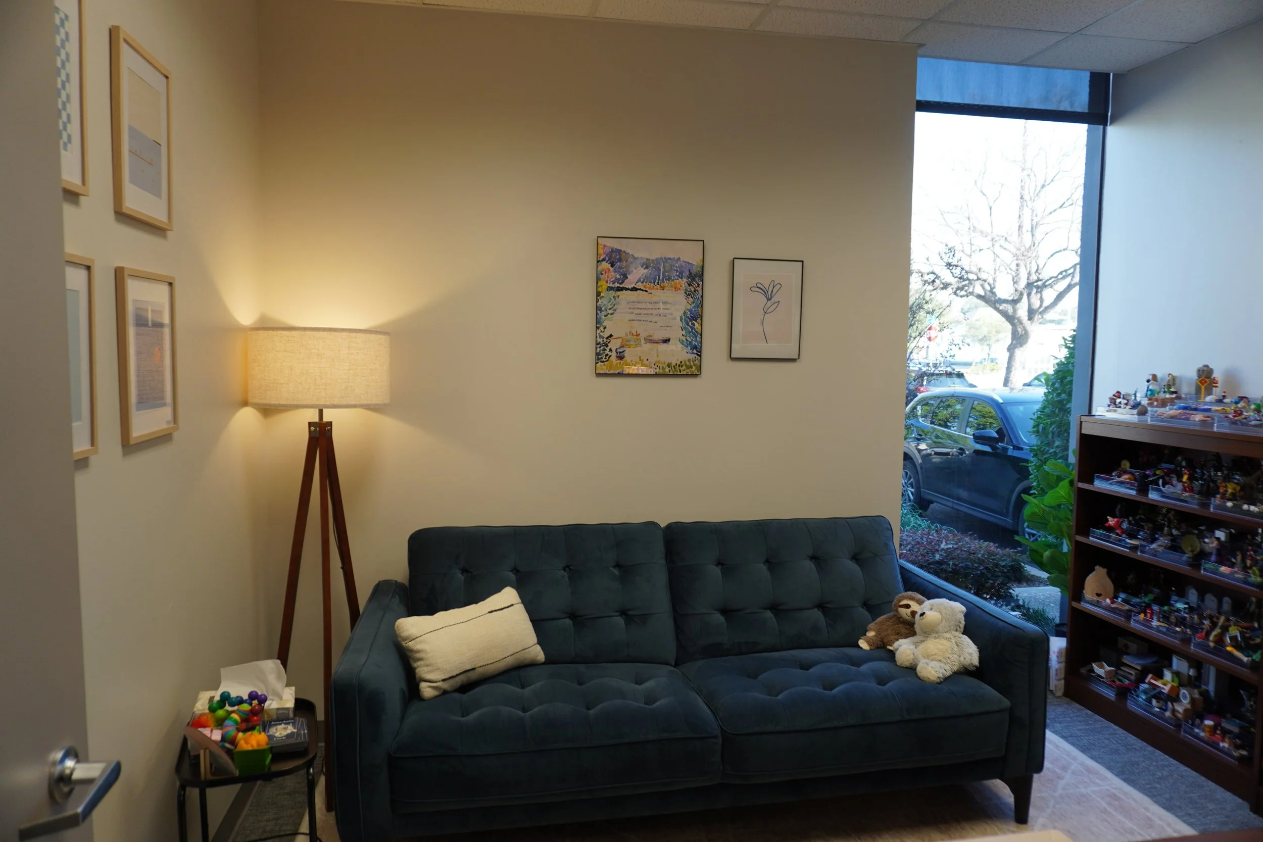 A cozy room features a blue tufted sofa with a pillow and plush toys. Soft lighting from a floor lamp and artwork adorn the walls, exuding a warm, inviting atmosphere.