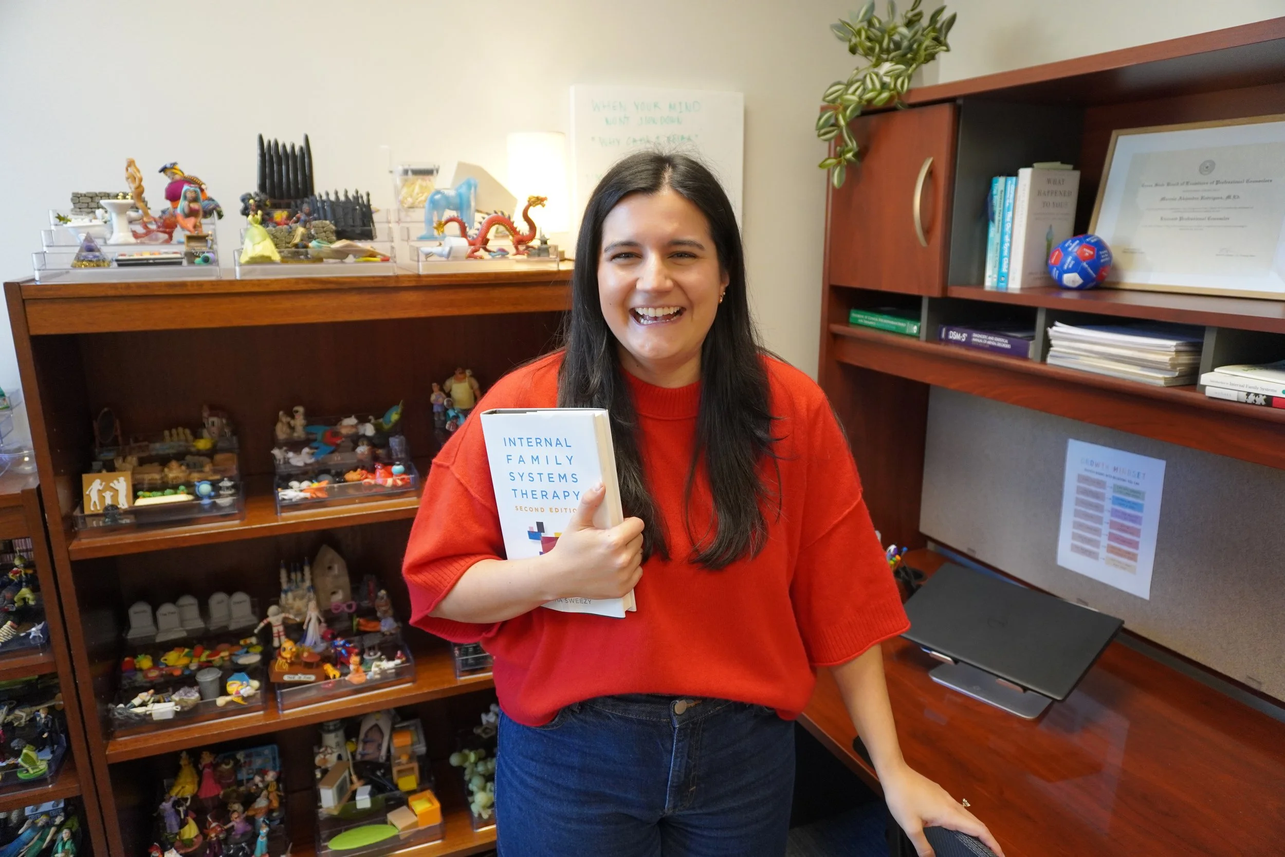Smiling woman in red holds a book in an office with a display of colorful figurines on shelves, conveying a cheerful and academic atmosphere.